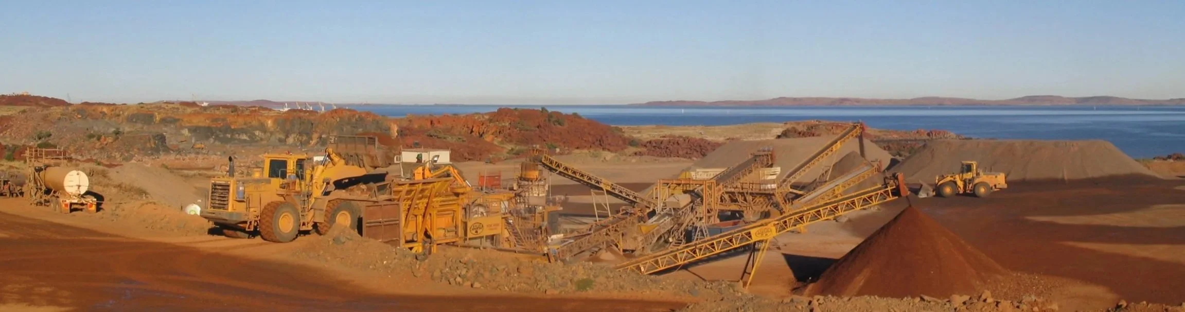 Open-pit mining site with large yellow machinery and conveyor belts, with a body of water and hills in the background.