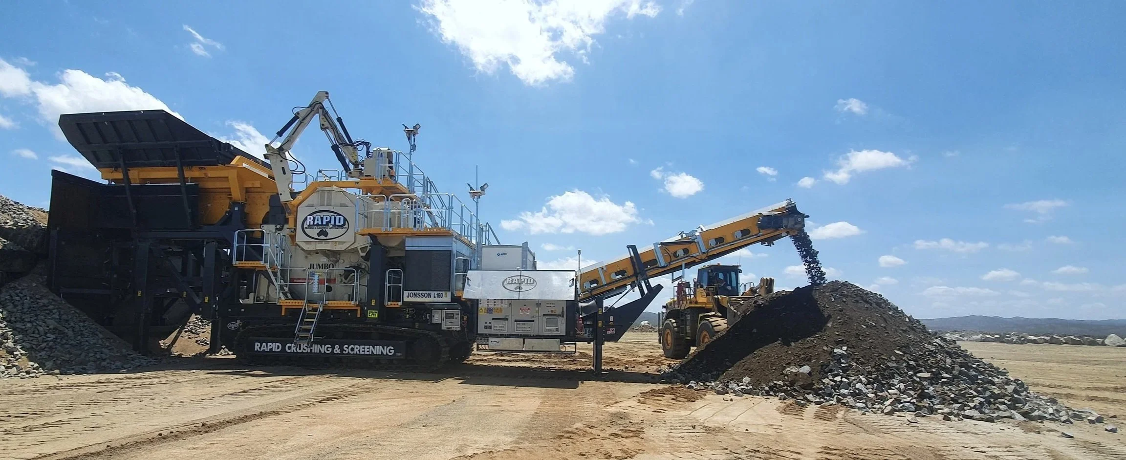 A large industrial rock crushing and screening machine operating outdoors, with a conveyor belt transferring crushed material into a heap, under a partly cloudy sky.