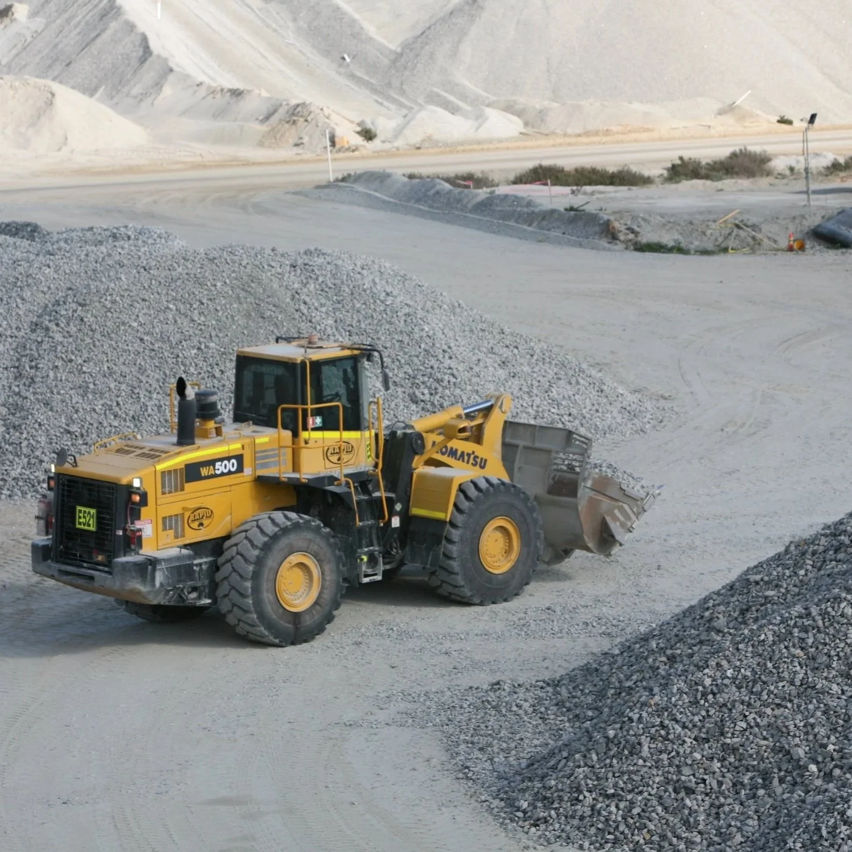 A yellow Komatsu WA500 wheel loader moving gravel in a construction or mining site with dirt and rocky terrain in the background.