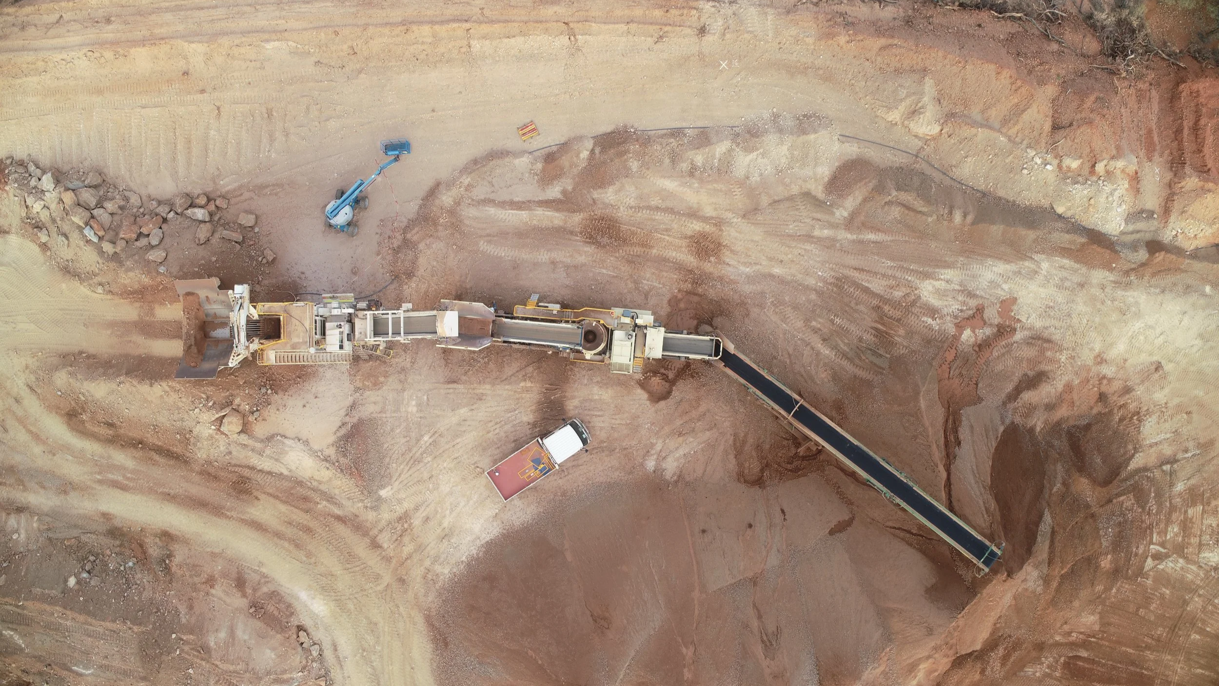 An aerial view of a construction site with large equipment, including a conveyor belt and a crane, on exposed earth and rocky terrain.