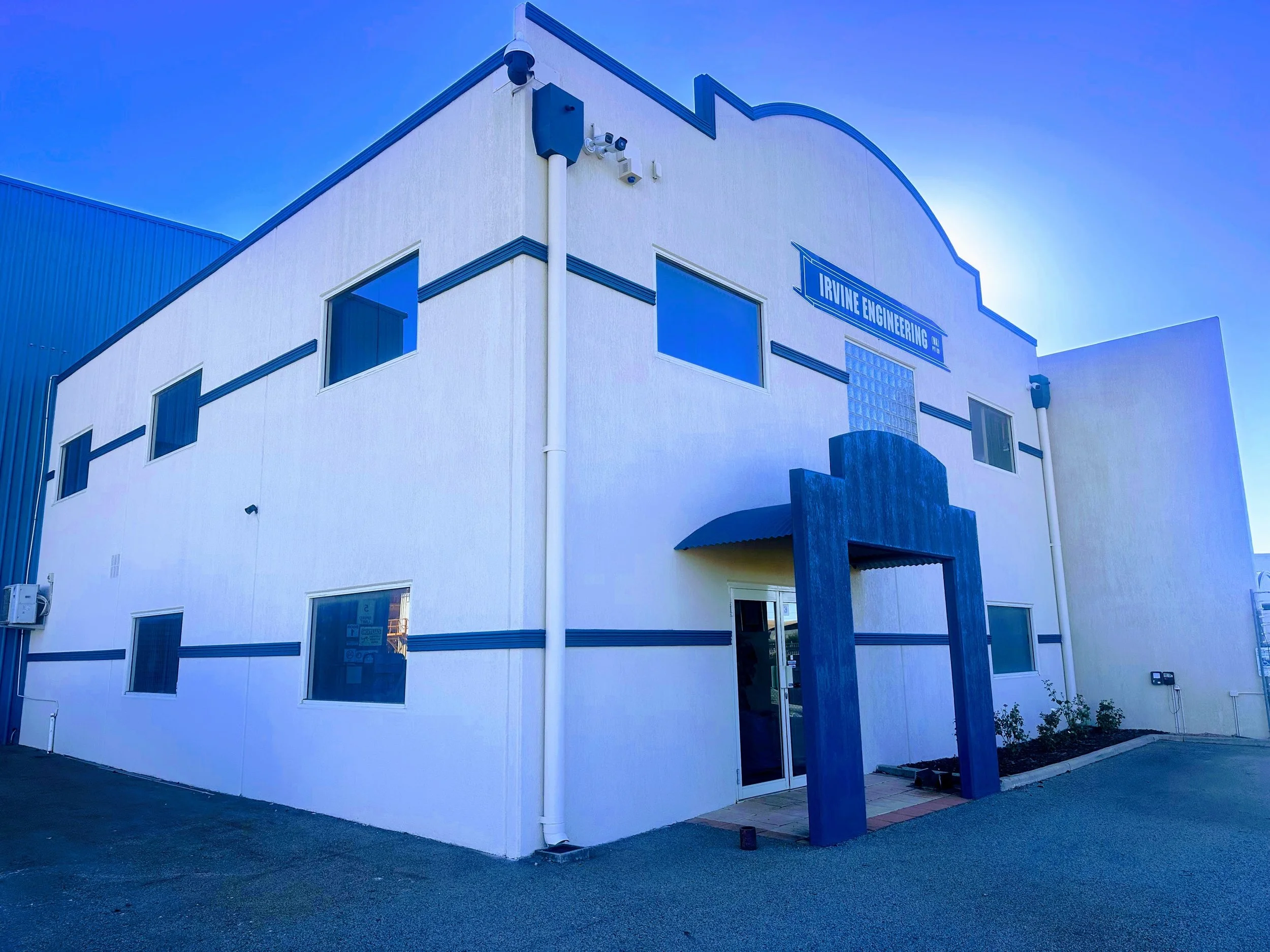A white two-story building with blue accents, labeled Irvine Engineering, featuring a decorative blue archway and multiple windows, with a blue building attached on the left and a small garden bed in front.