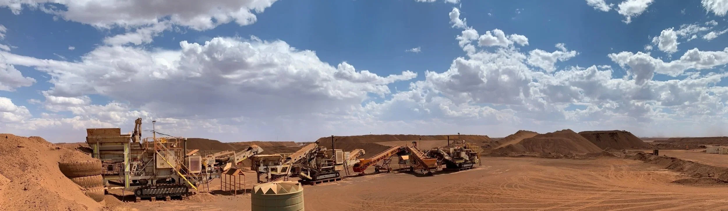 An expansive view of a mining site with multiple large industrial machines and conveyor belts in a desert landscape under a cloudy sky.