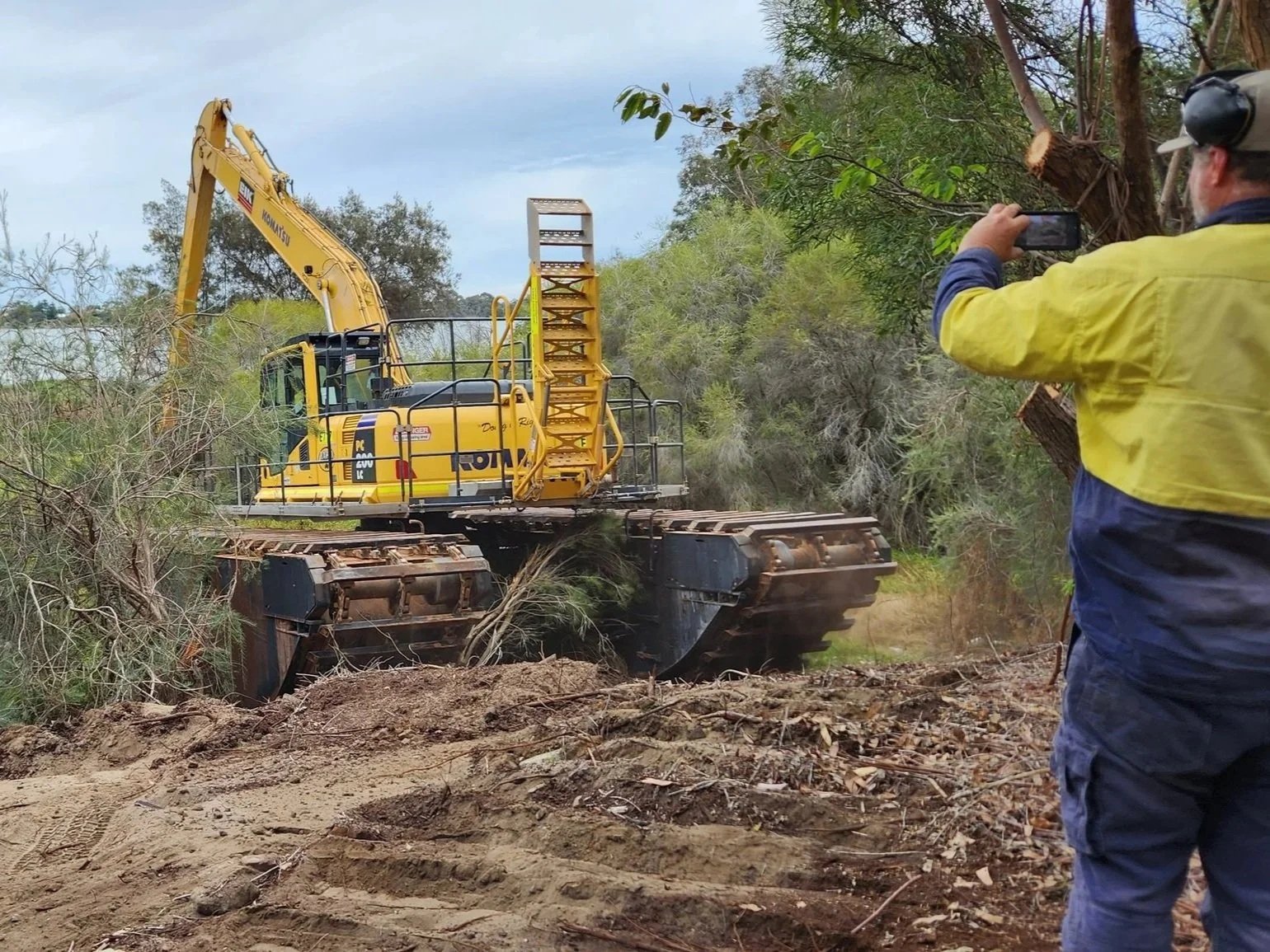 A person in a yellow jacket is taking a photo of a yellow excavator working on a dirt and tree clearing project in a wooded area.
