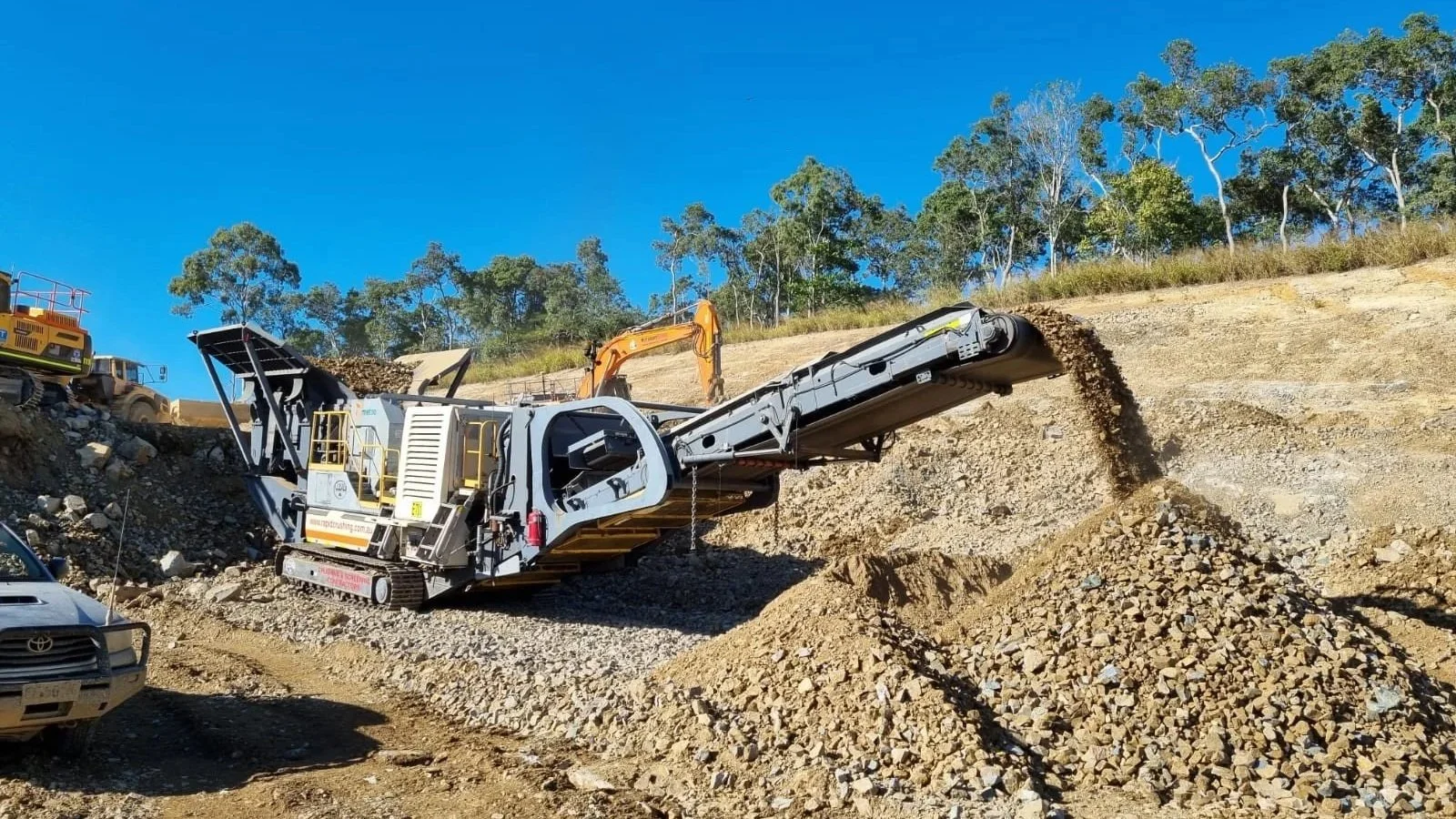 Large gravel screener machine at a construction site with gravel and dirt, surrounded by construction vehicles, under a clear blue sky and trees in the background.