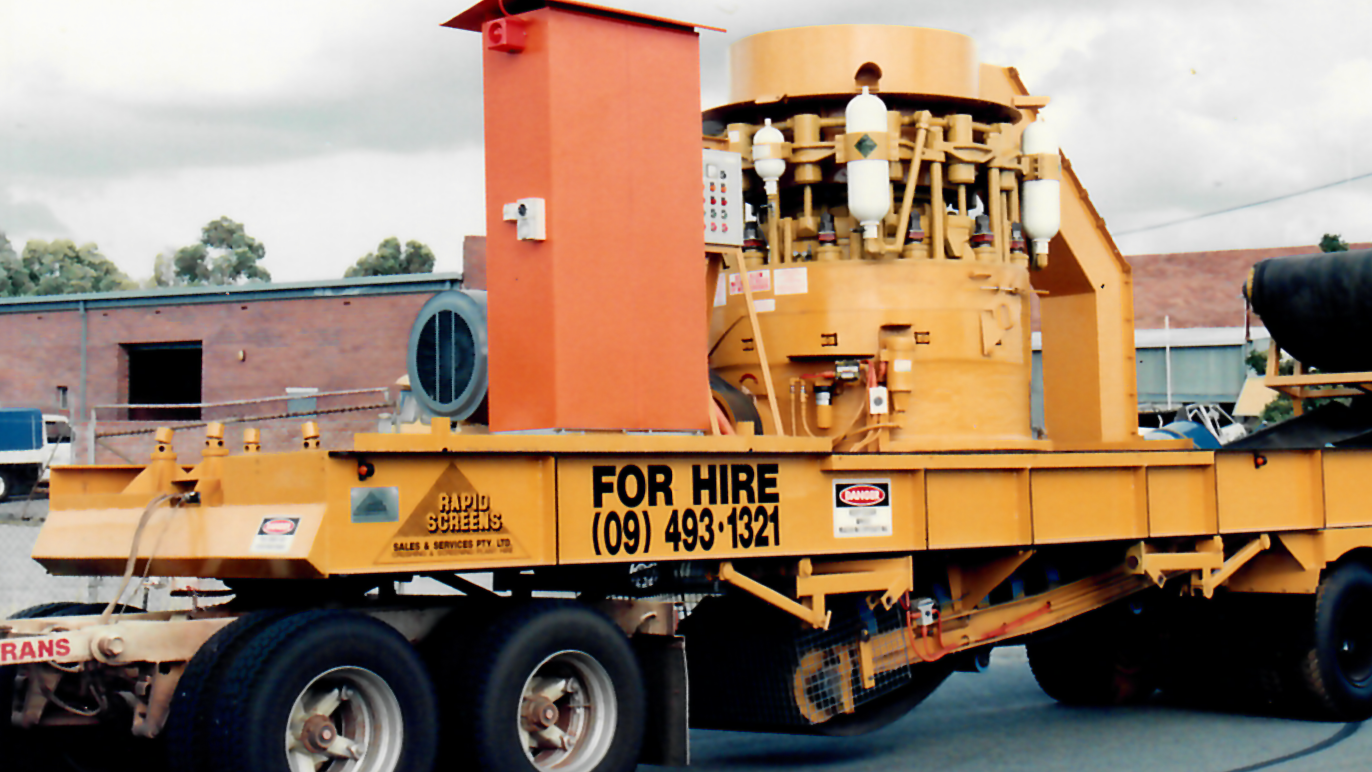 A large yellow industrial machine on a trailer, with a sign that says "FOR HIRE" and a phone number, parked outdoors near a brick building.