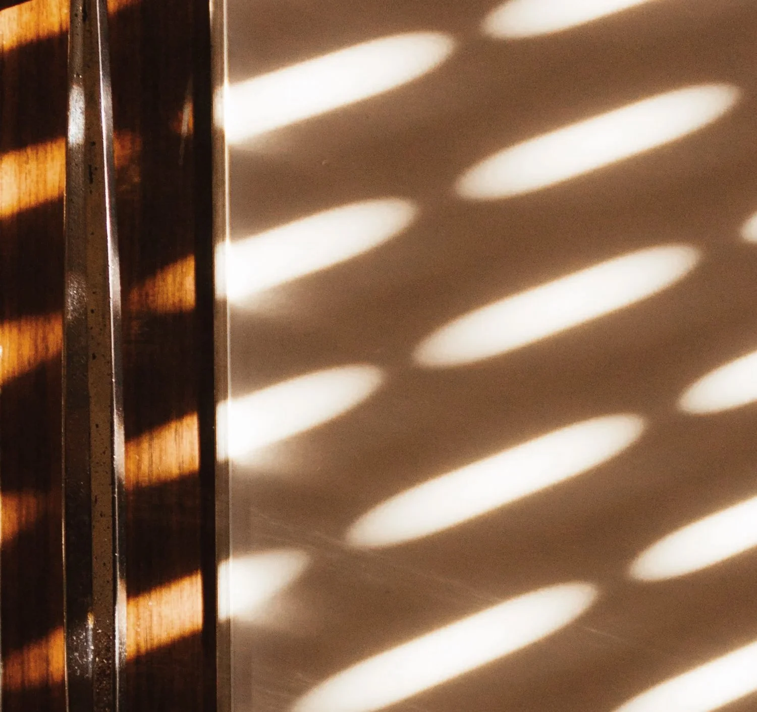 Sunlight casts diagonal shadows through a striped slatted surface onto a wall, creating an abstract pattern.