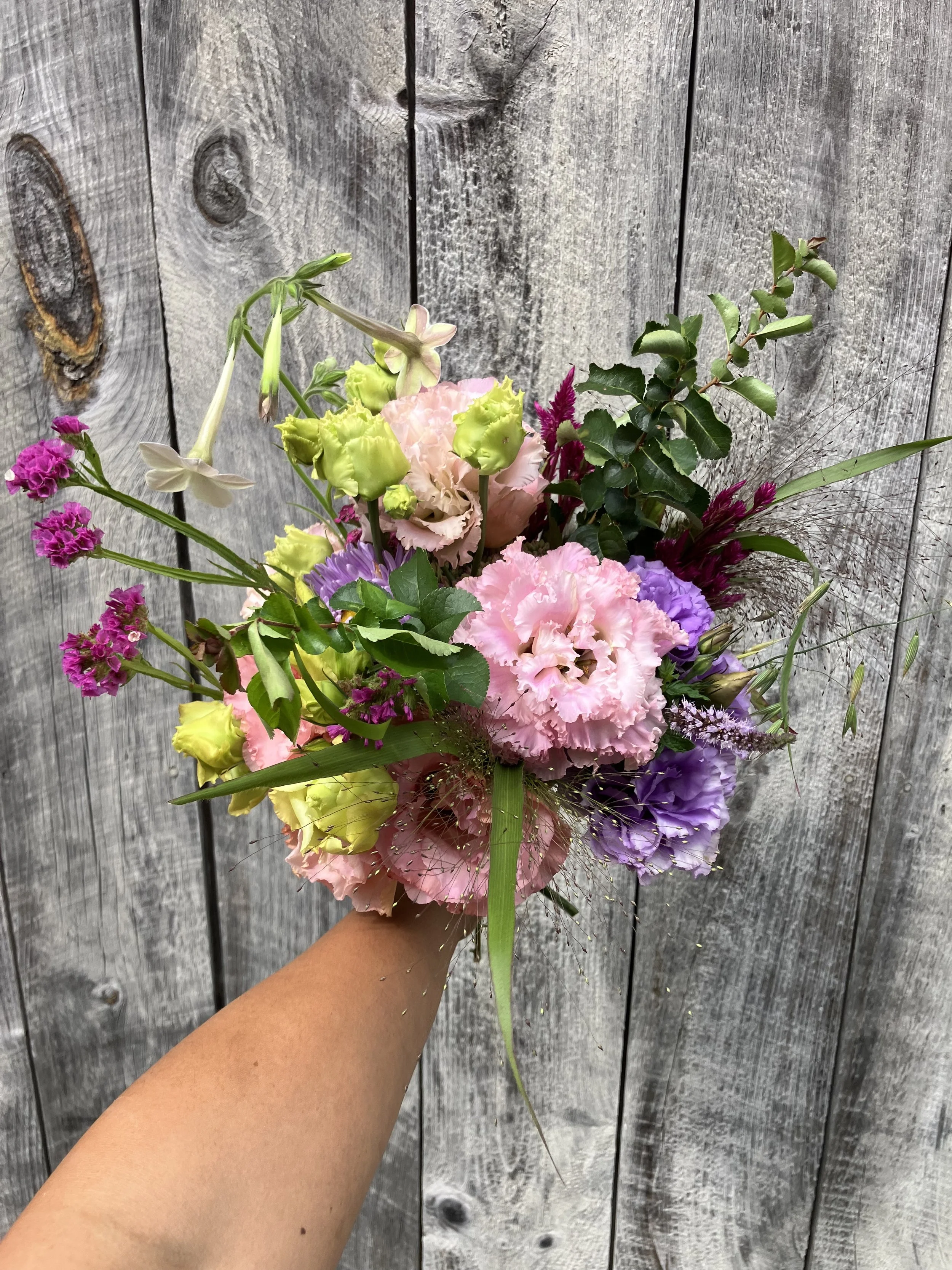 A bridesmaid or attendant bouquet is held in front of rustic barn board. It contains lisanthus, nicotiana, and lots of texture and whimsy. 