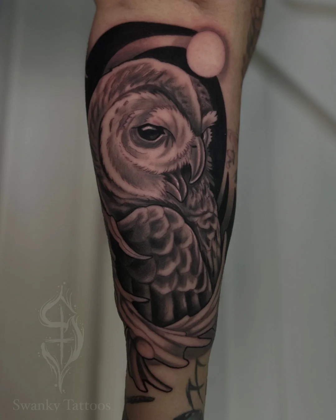 Black and gray tattoo of an owl on a person's arm, featuring detailed feathers and a large, intense eye, with a moon and abstract background elements.