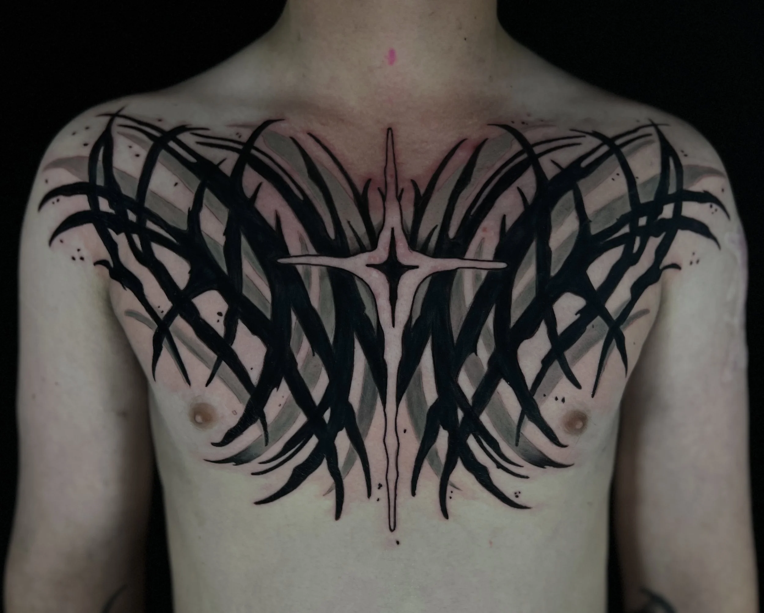 Black tribal-style chest tattoo design with sharp lines and symmetrical patterns on person's chest and shoulders.