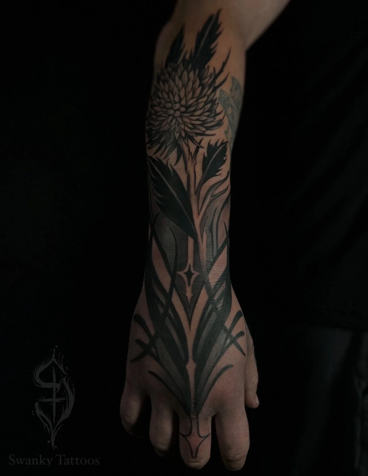 A black ink tattoo on a person's forearm and hand, featuring a floral design with sharp, geometric elements.