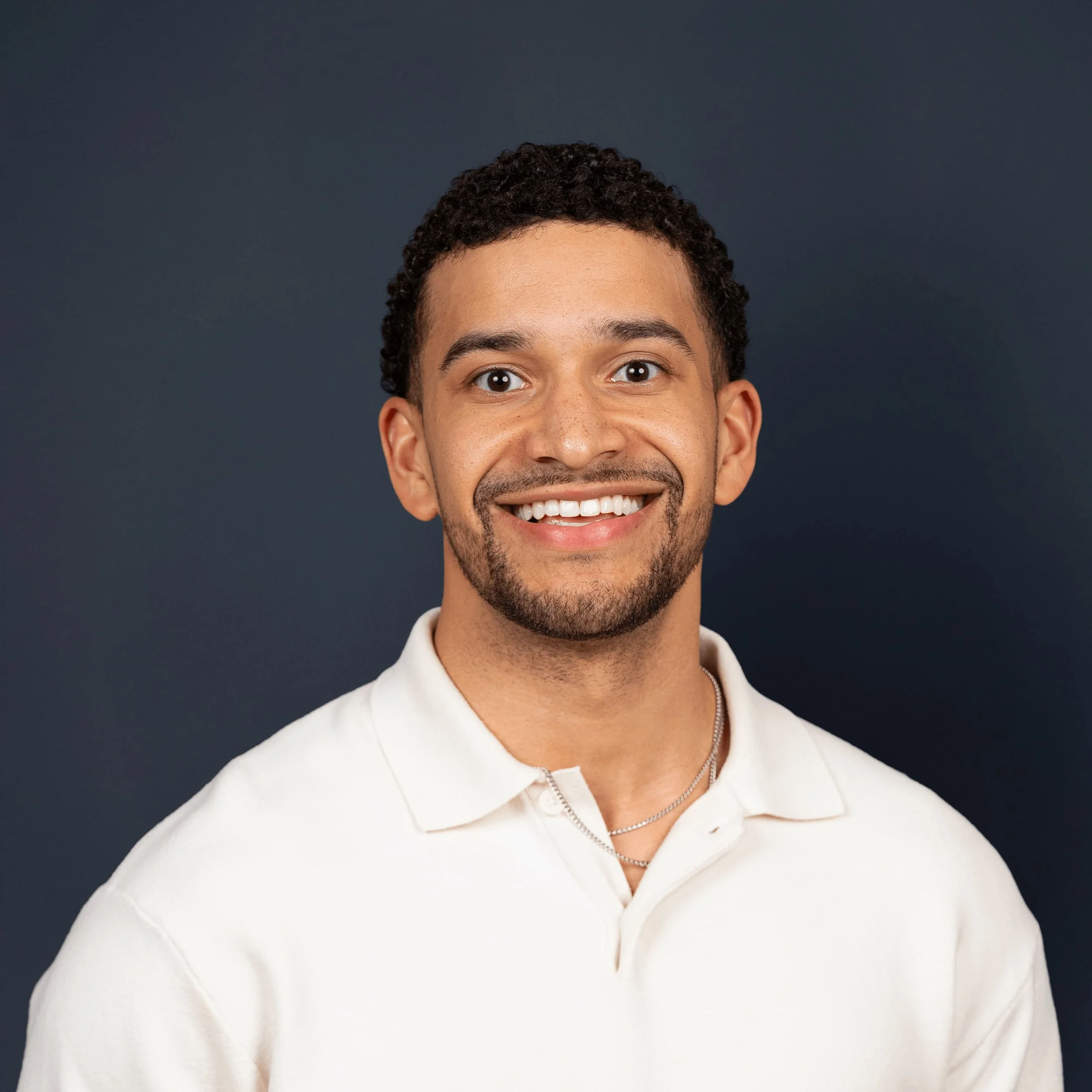 Smiling young man with short curly hair, a beard, and a white polo shirt against a dark background.