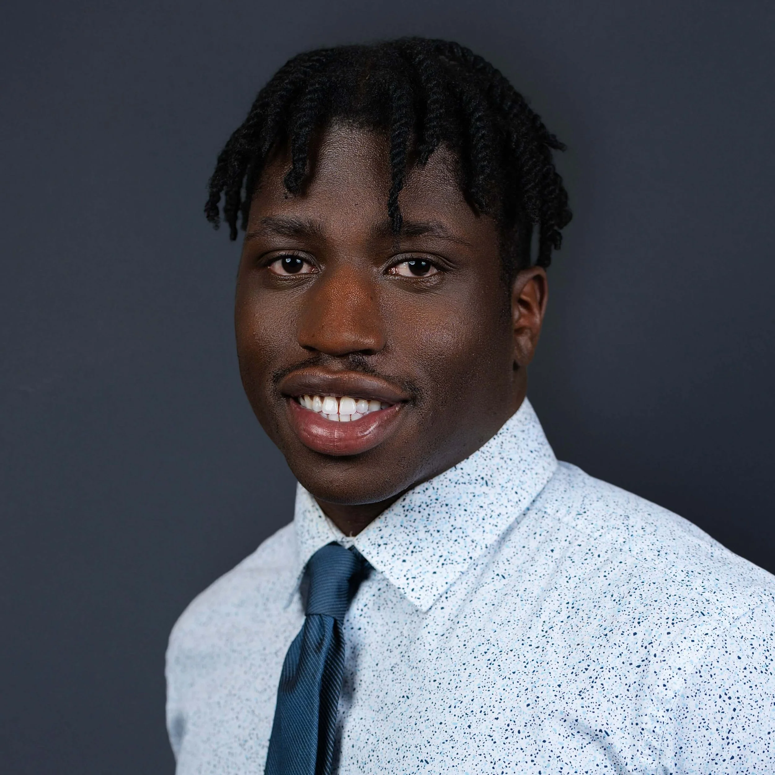 Headshot of a young man with dark skin, styled dreadlocks, wearing a white shirt with a navy tie, smiling slightly, against a dark background.