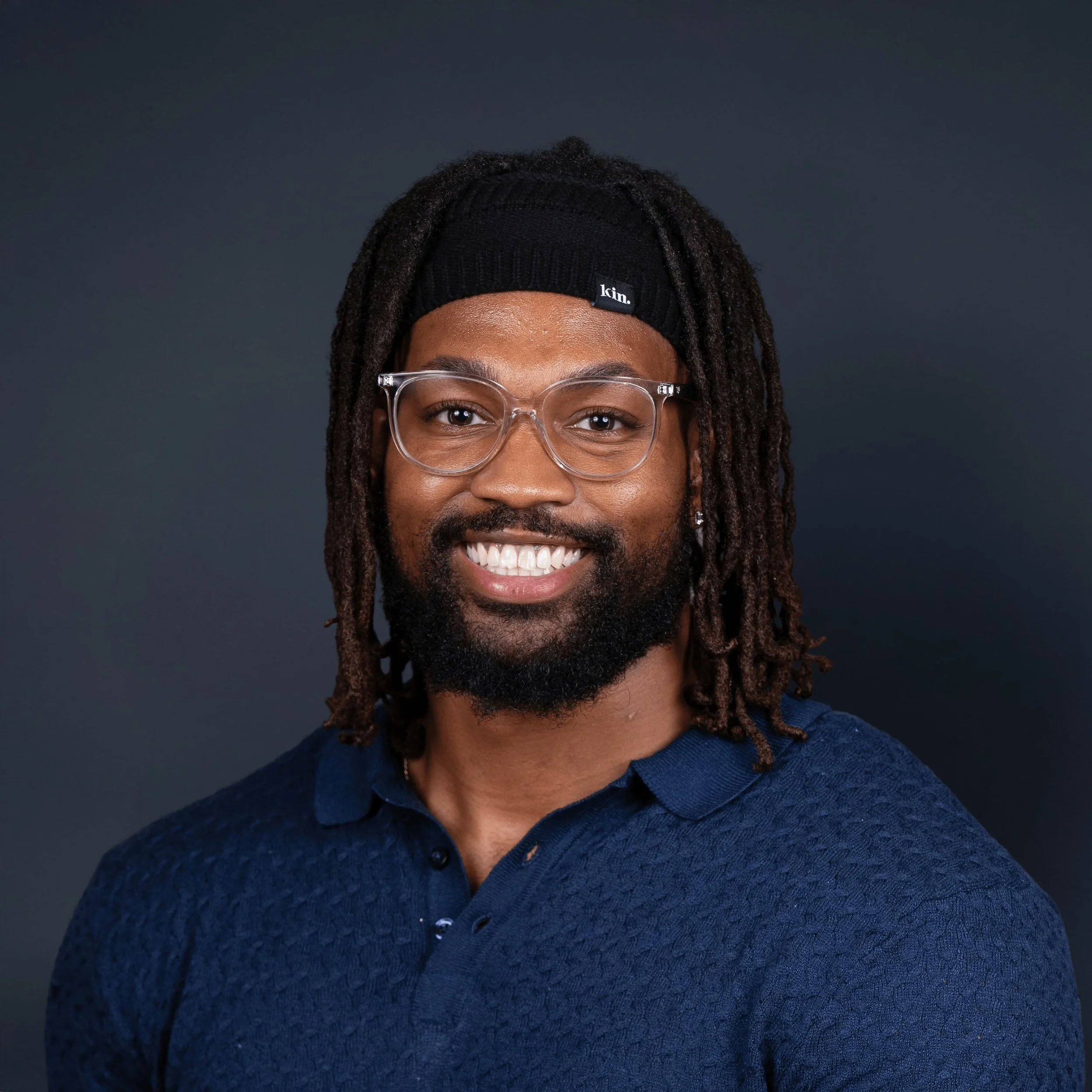 Headshot of a smiling man with dreadlocks, wearing glasses, a black knit beanie, and a navy blue polo shirt, against a dark background.