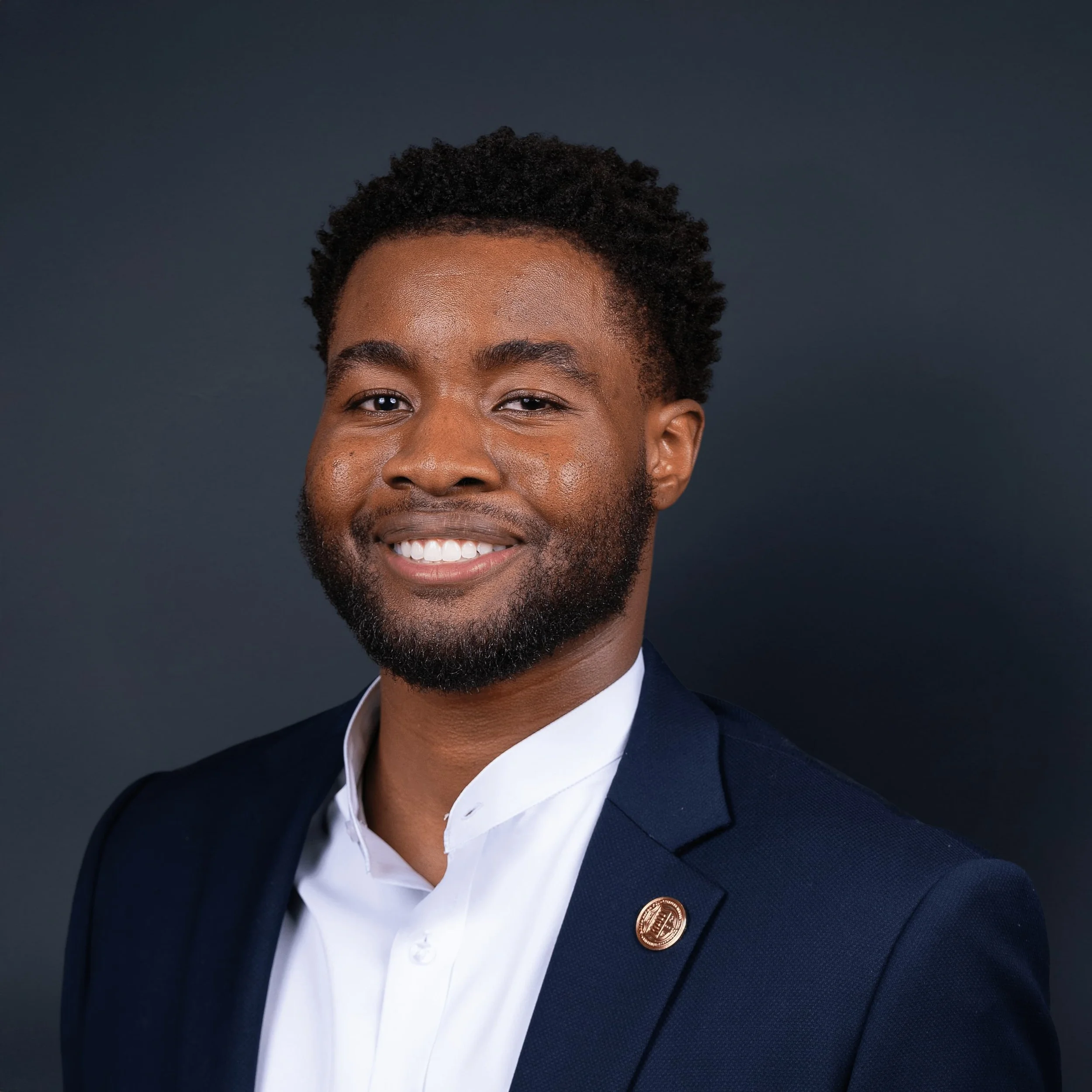 A smiling man with a beard wearing a navy blazer with a lapel pin and a white shirt against a dark background.