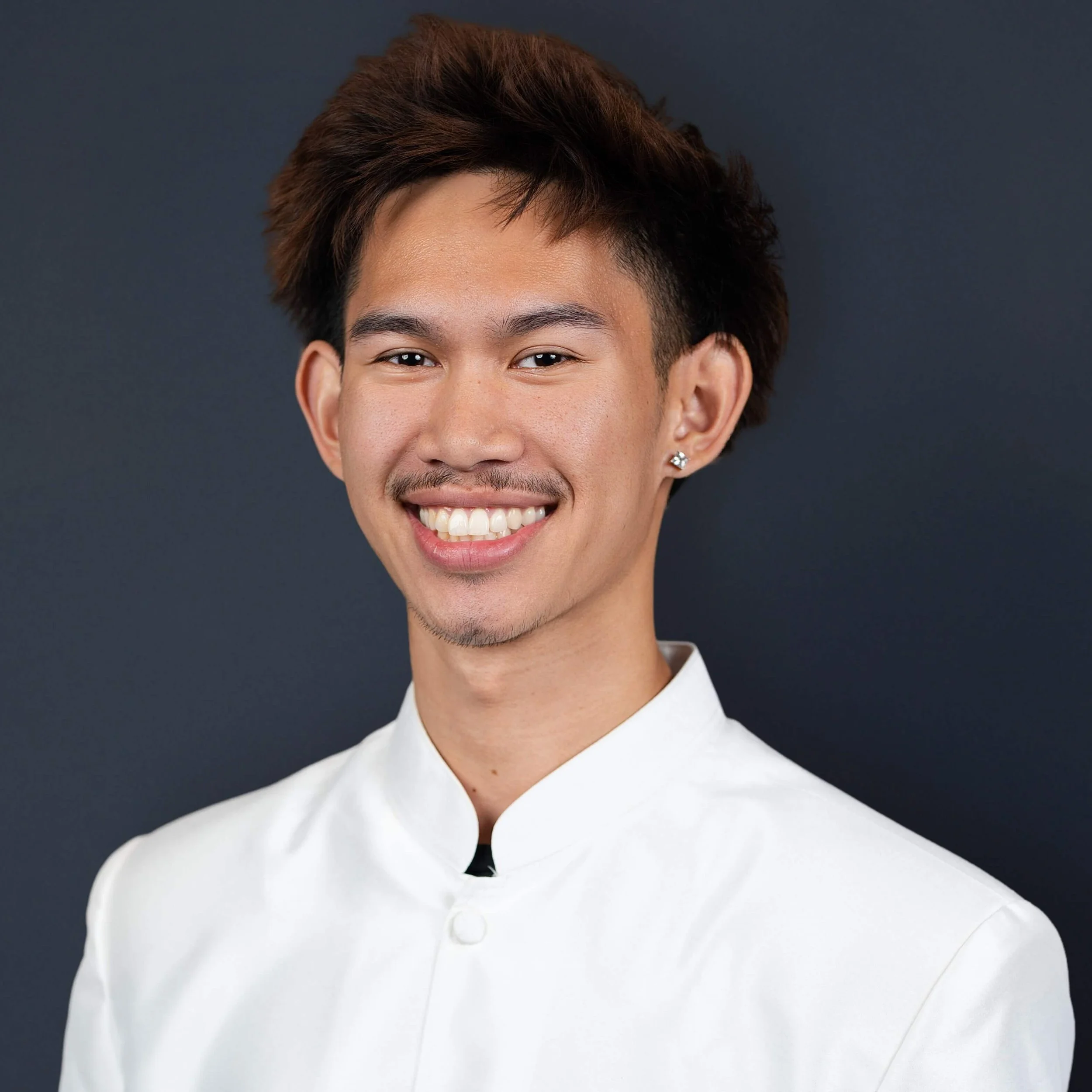 A young man with brown hair, a mustache, and a goatee, smiling, wearing a white chef's jacket against a dark background.