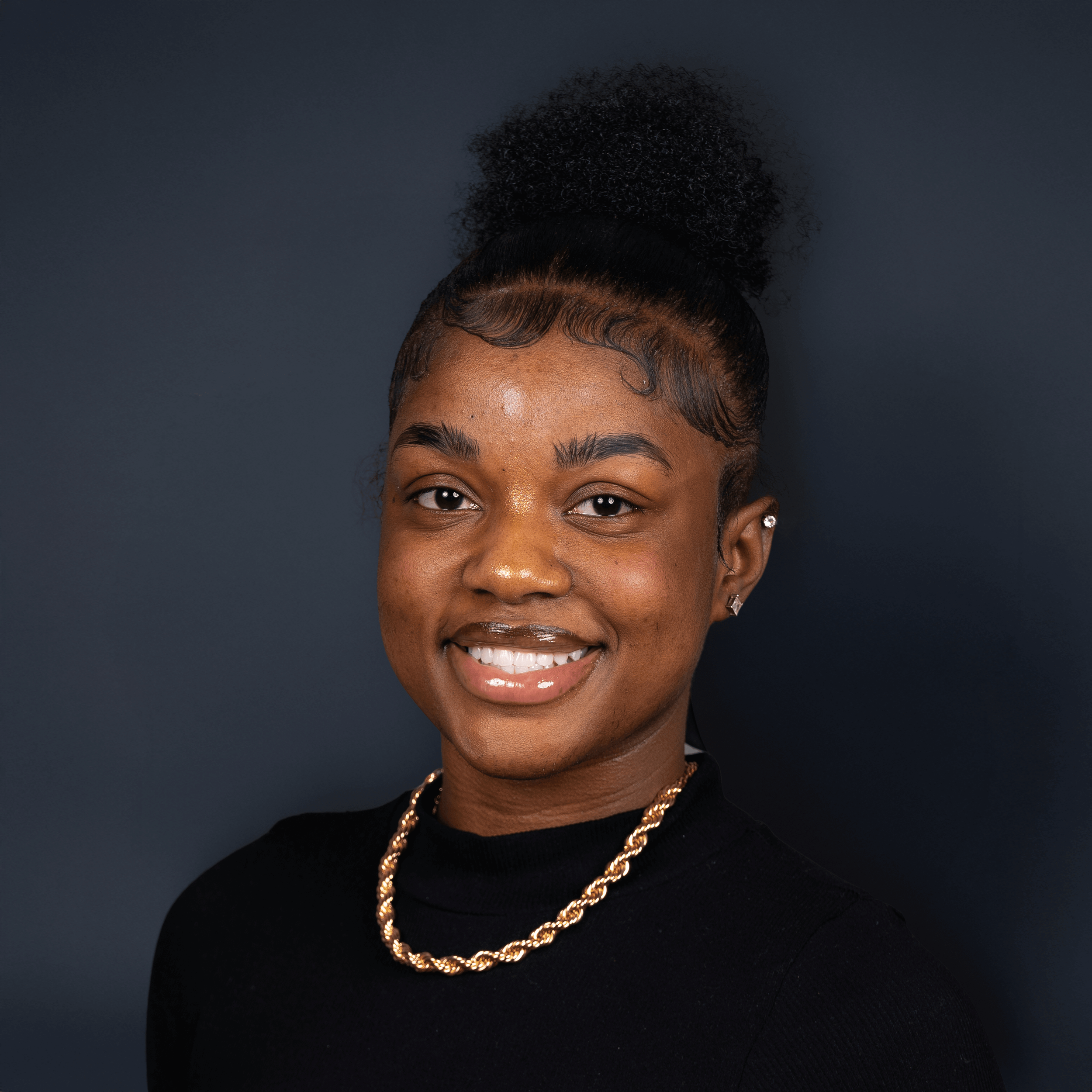 Portrait of a young woman smiling against a dark blue background, wearing a black top, gold chain necklace, and small earrings, with her hair styled in a high curly bun.