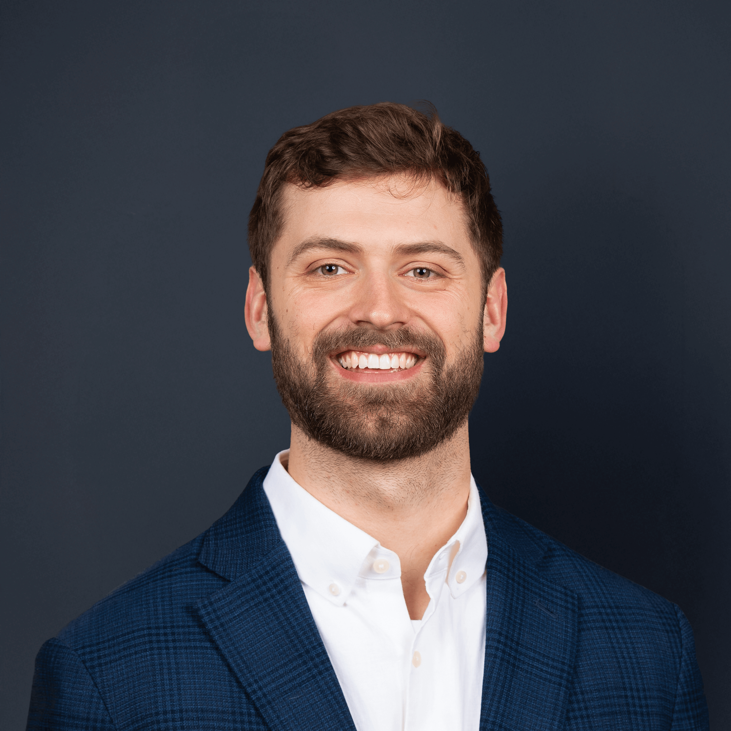 Professional portrait of a smiling man with brown hair and beard, wearing a blue checkered blazer and white shirt, against a dark background.