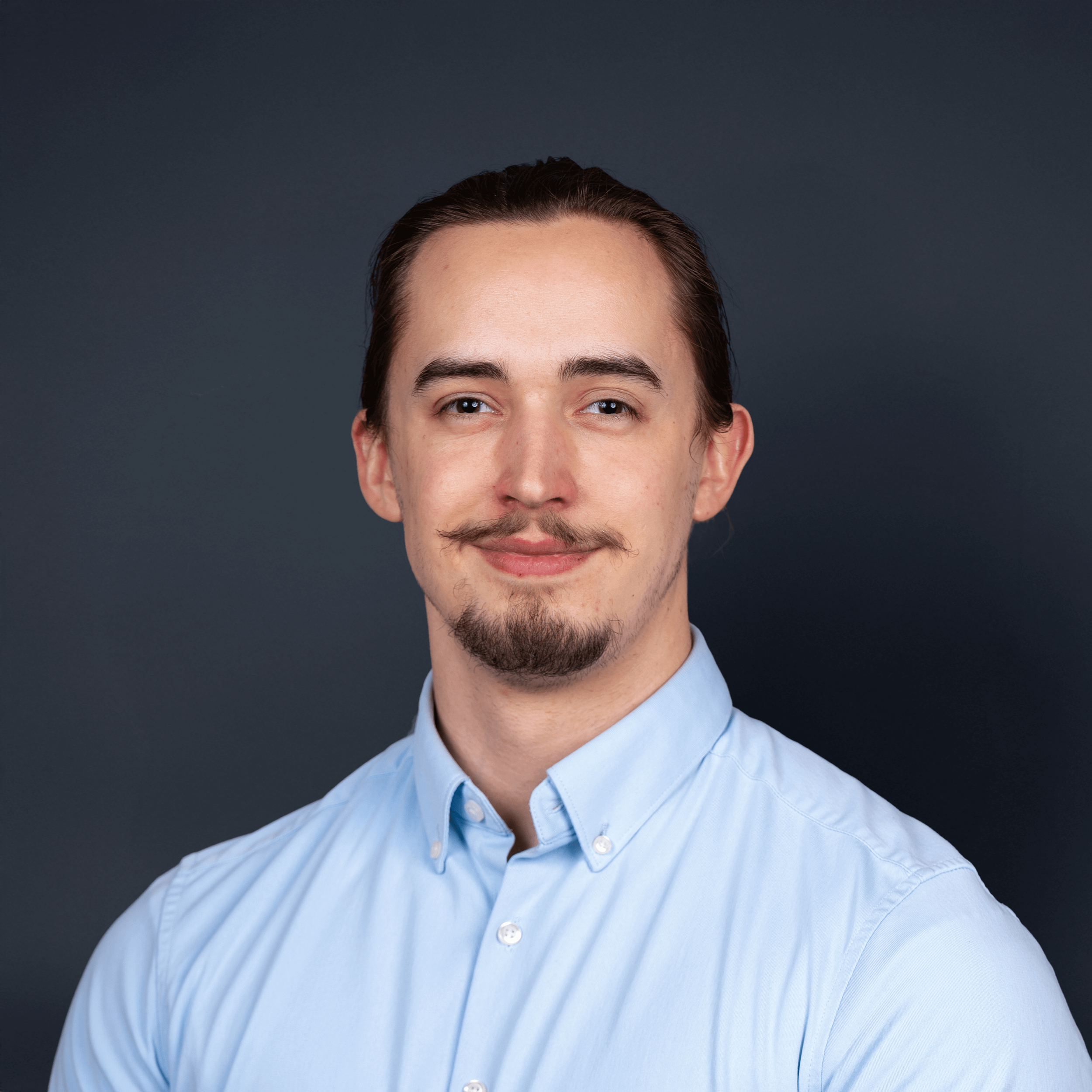 A portrait of a young man with light skin, dark brown hair, and a mustache and goatee, wearing a light blue button-up shirt, smiling softly, against a dark background.