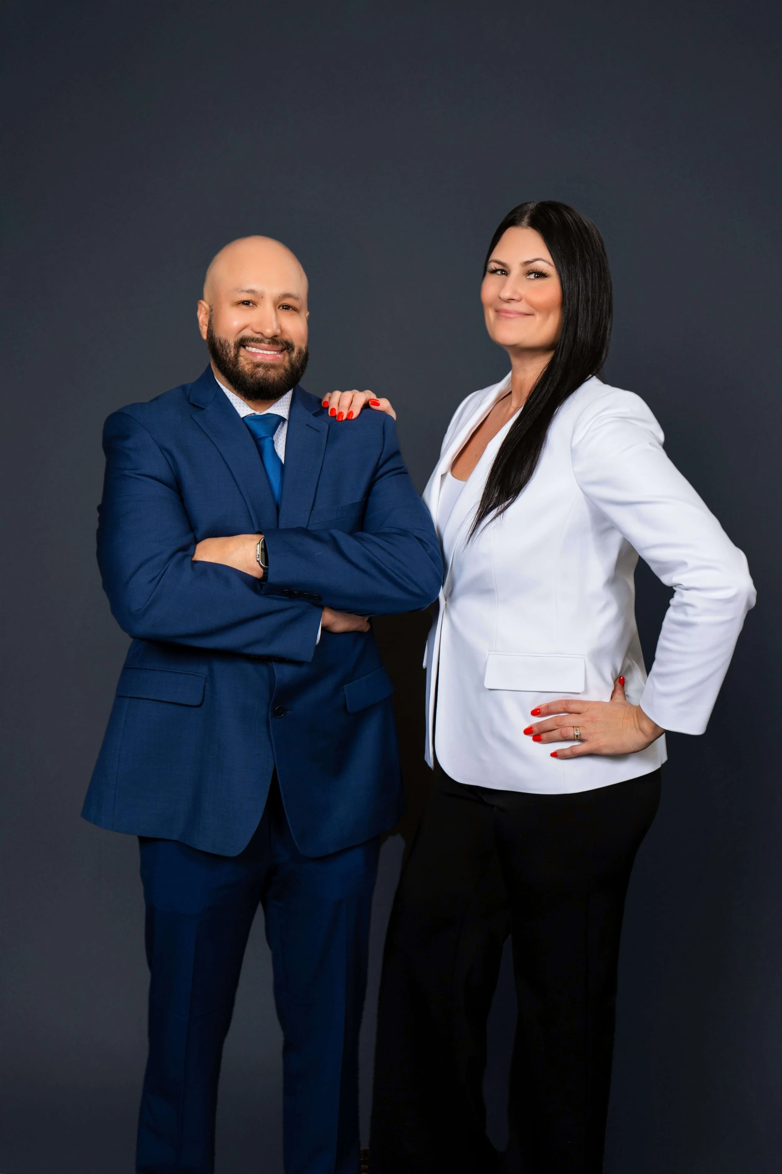 A man and a woman in professional attire standing against a dark background. The man is wearing a blue suit and tie, and the woman is wearing a white blazer with black pants. They are smiling and posing confidently.