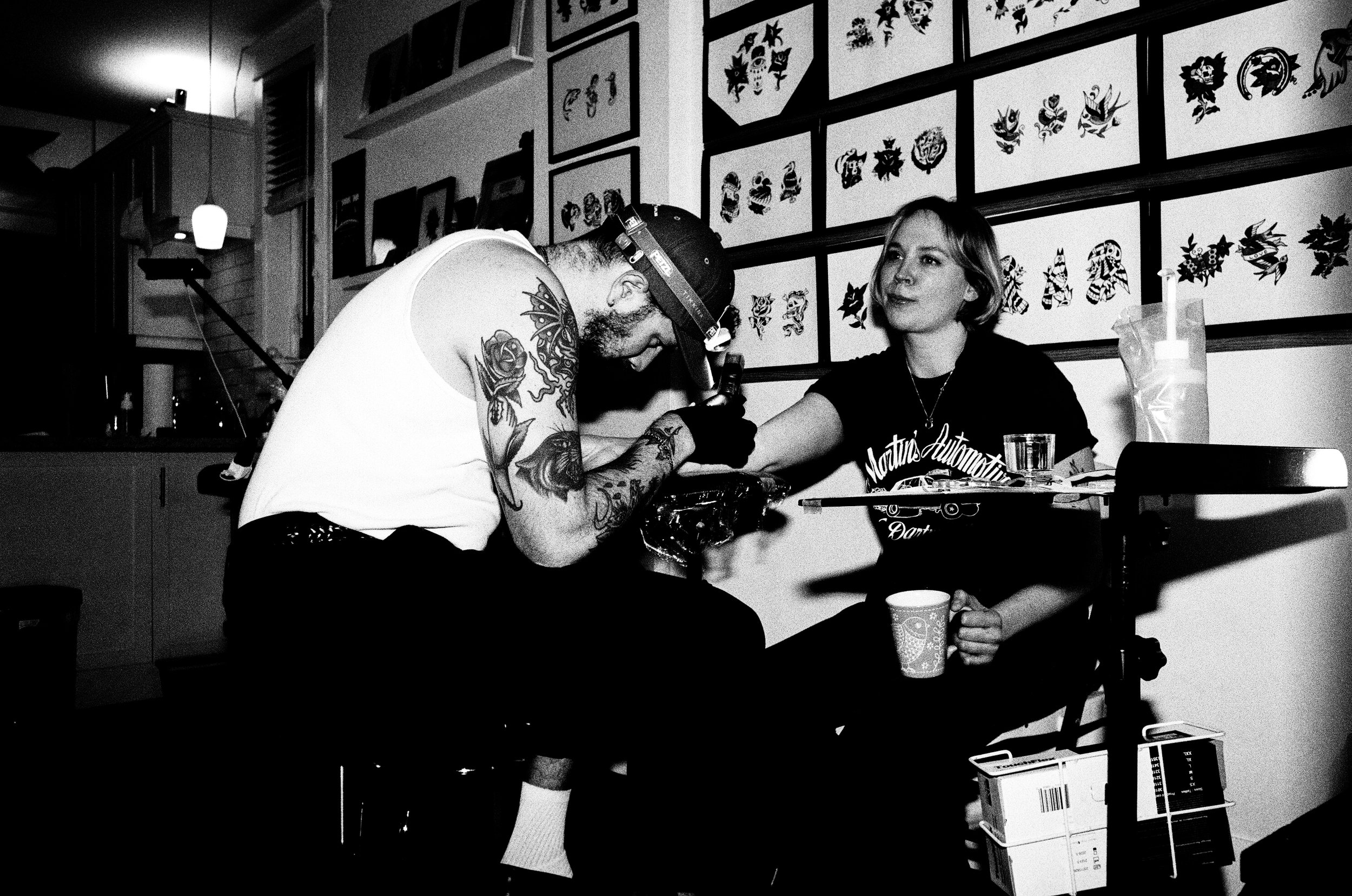 A tattoo artist tattooing a person's arm in a tattoo studio with framed artwork on the wall, while a woman sits nearby holding a cup.