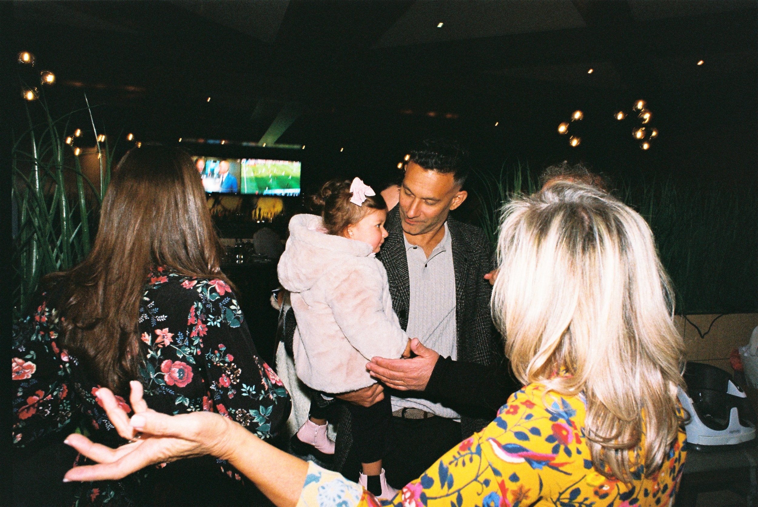 A group of people, including a man holding a young girl with a pink bow in her hair, are gathered indoors at a social event, with a television in the background showing a sports game.