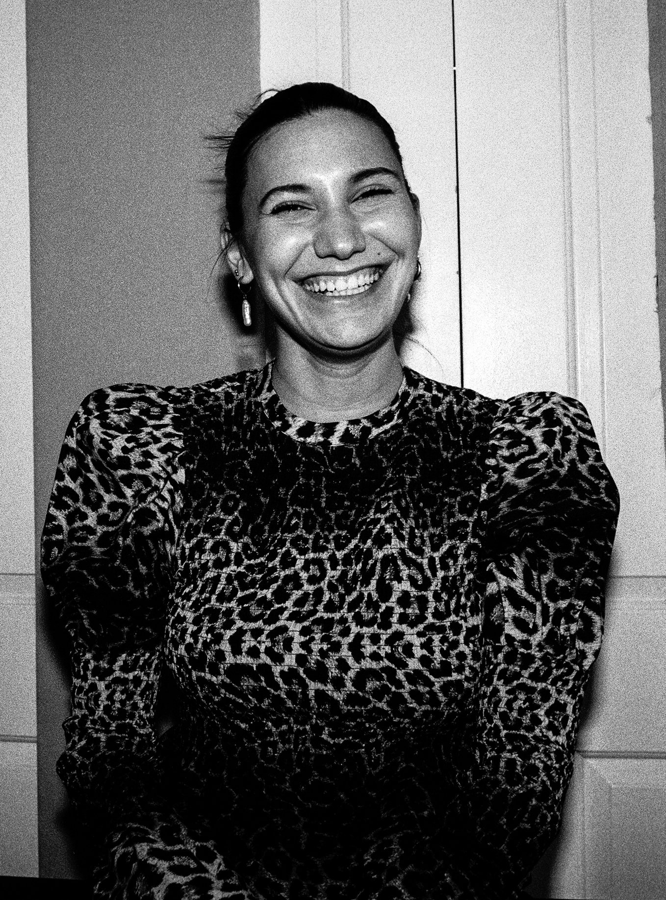 A woman with earrings smiling broadly in a black and white photo, wearing a leopard print blouse, standing in front of a door.
