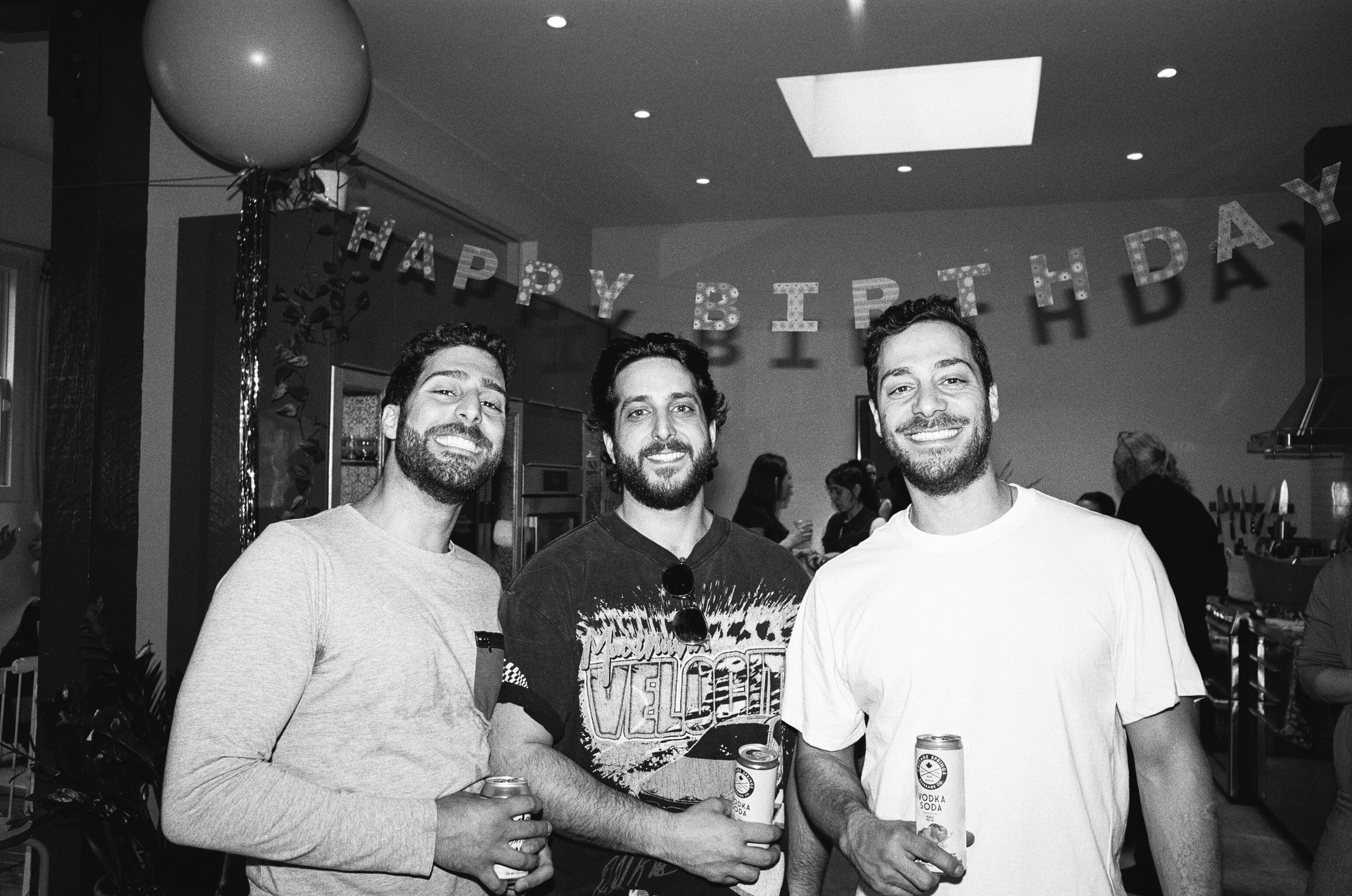 Three men smiling and holding cans of soda at a birthday party, with a "Happy Birthday" banner in the background.