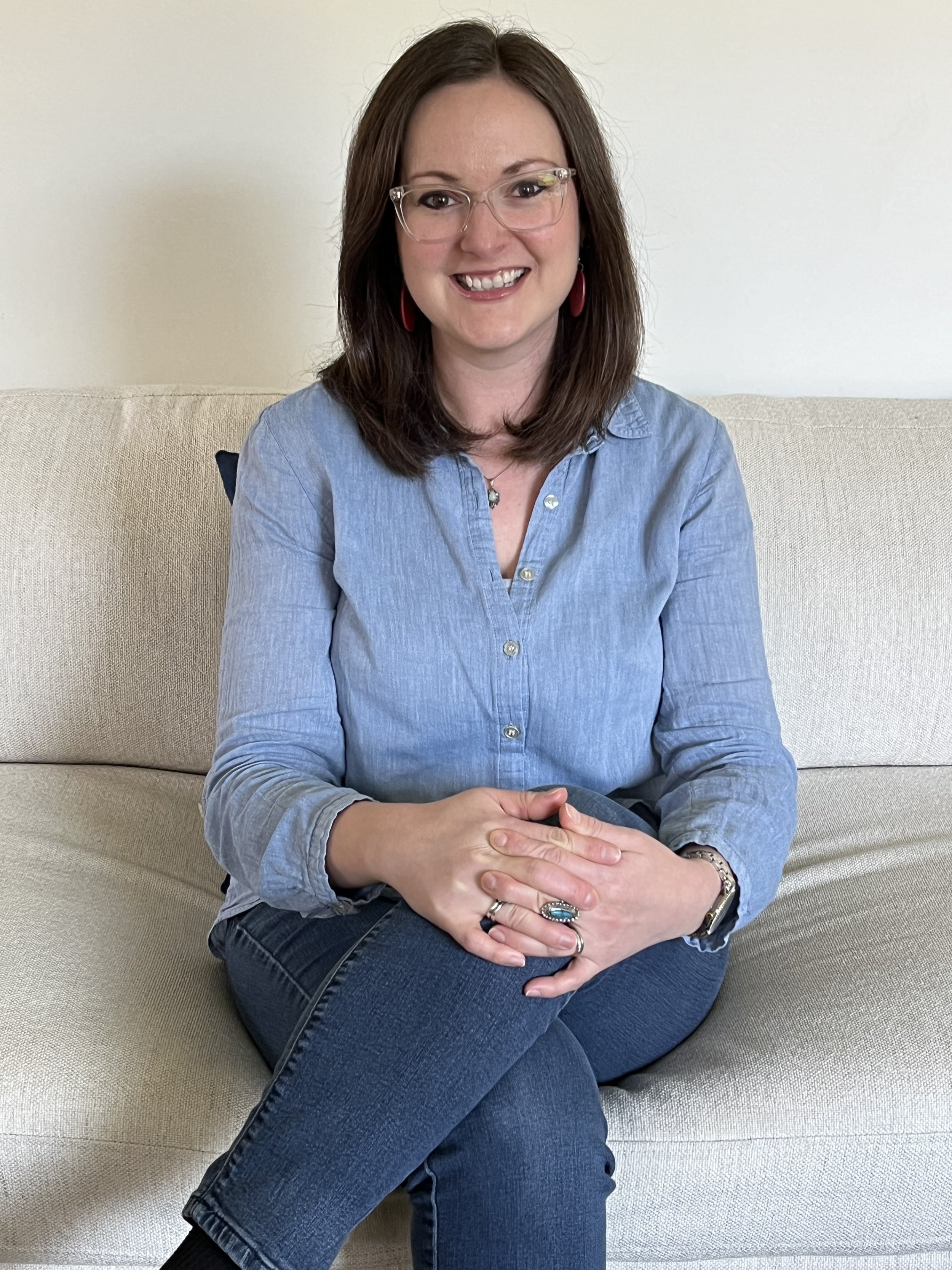 A woman with brown hair, glasses, and red earrings sitting on a beige sofa, smiling, wearing a light blue button-up shirt and jeans.