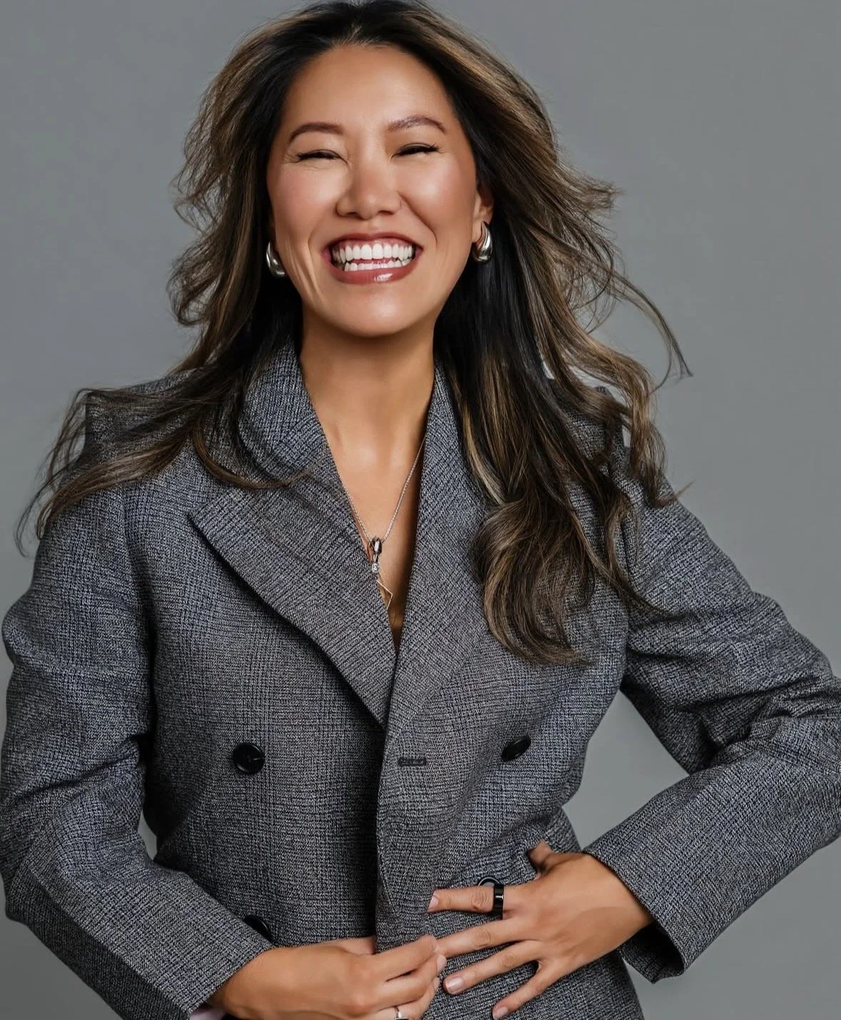 A headshot of a smiling woman with shoulder-length brown hair, wearing a gray blazer, jewelry, and a ring, against a plain gray background.