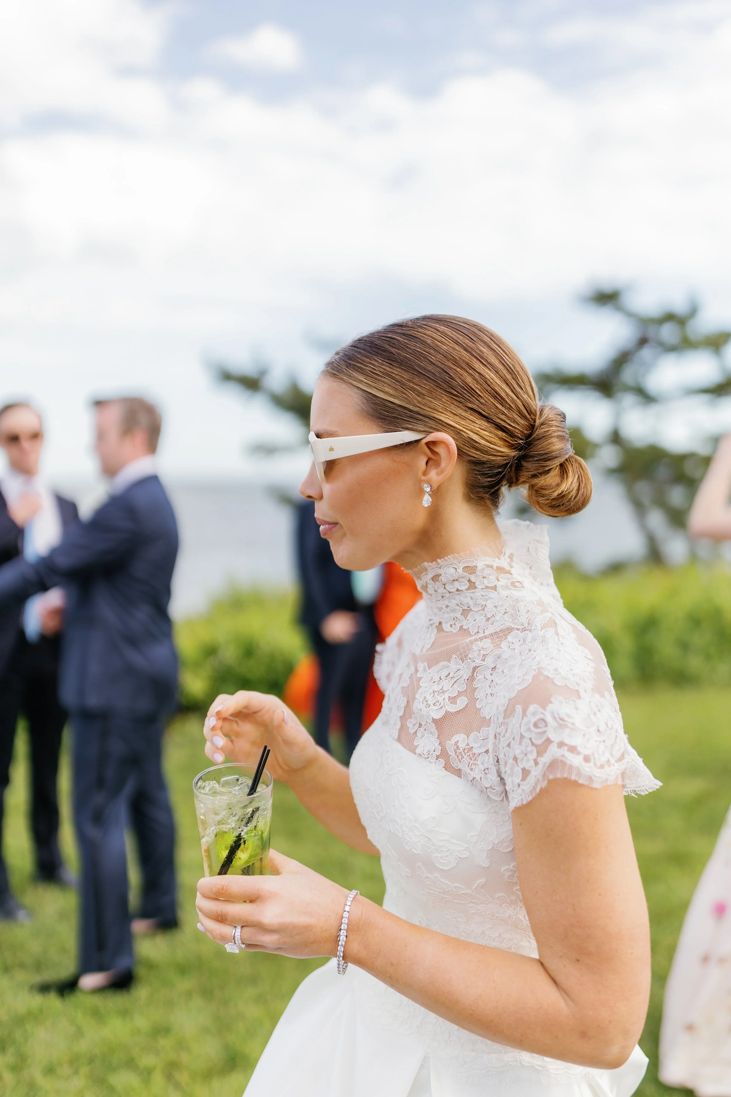 A bride in a white lace dress with her hair in a low bun, holding a glass of mint mojito, at an outdoor gathering with blurred background of people in suits and a cloudy sky.