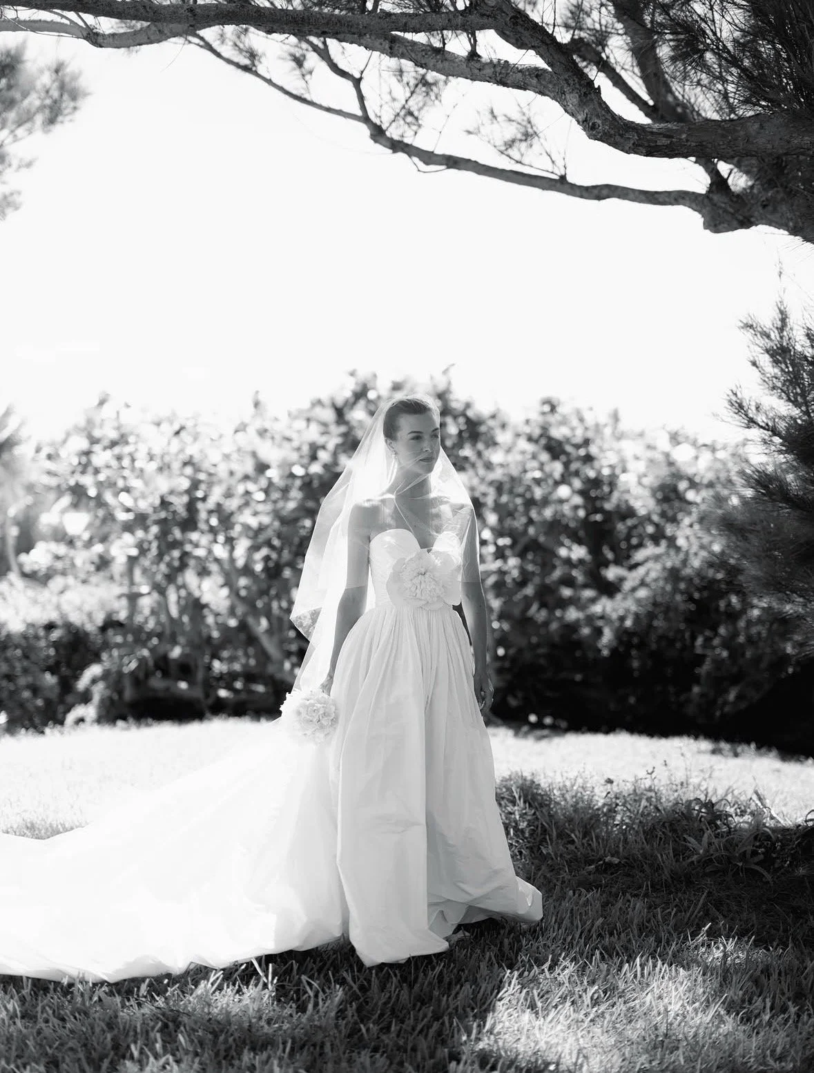 A bride in a strapless wedding gown with a full skirt and veil, standing outdoors on grass with trees and shrubs in the background, in black and white.