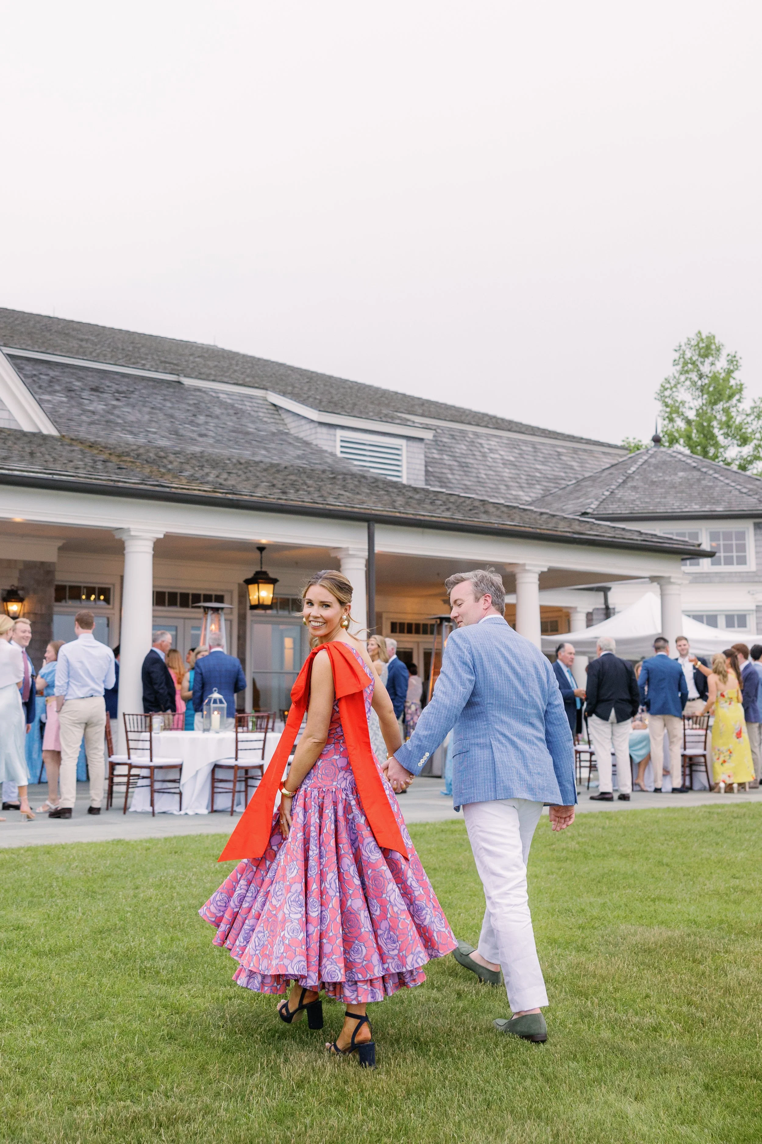A couple holding hands and dancing on a lawn at an outdoor wedding reception, with guests and a large house with a porch in the background.