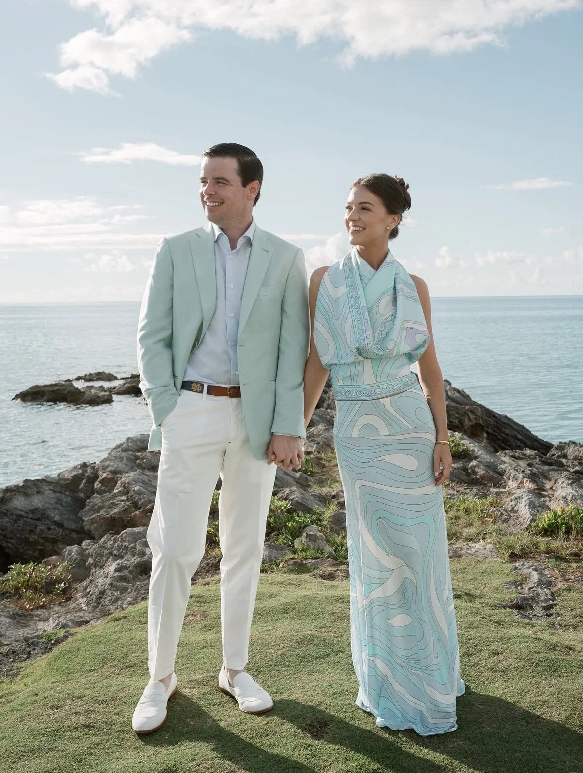 A couple holding hands stands on a grassy area near rocks, with the ocean and a partly cloudy sky in the background, dressed in light, pastel-colored clothes ahead of their welcome party.