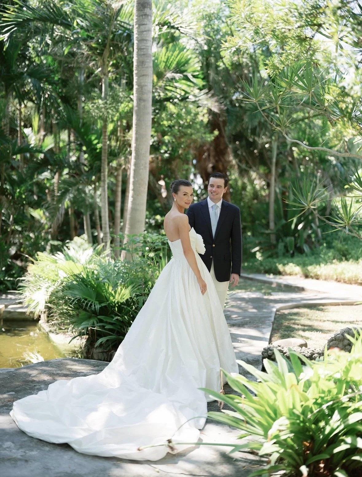 A bride and groom standing outdoors in a lush tropical garden. The bride is in a white strapless wedding gown with a long train, and the groom is in a dark suit with a white shirt and light tie. They are smiling, surrounded by green plants and tall p