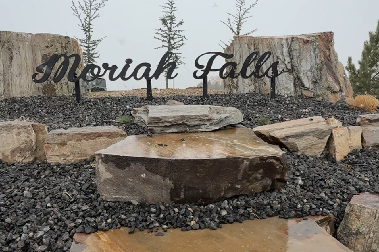 Sign reading 'Morial Falls' positioned among large rocks and pebbles at an outdoor location with trees in the background.