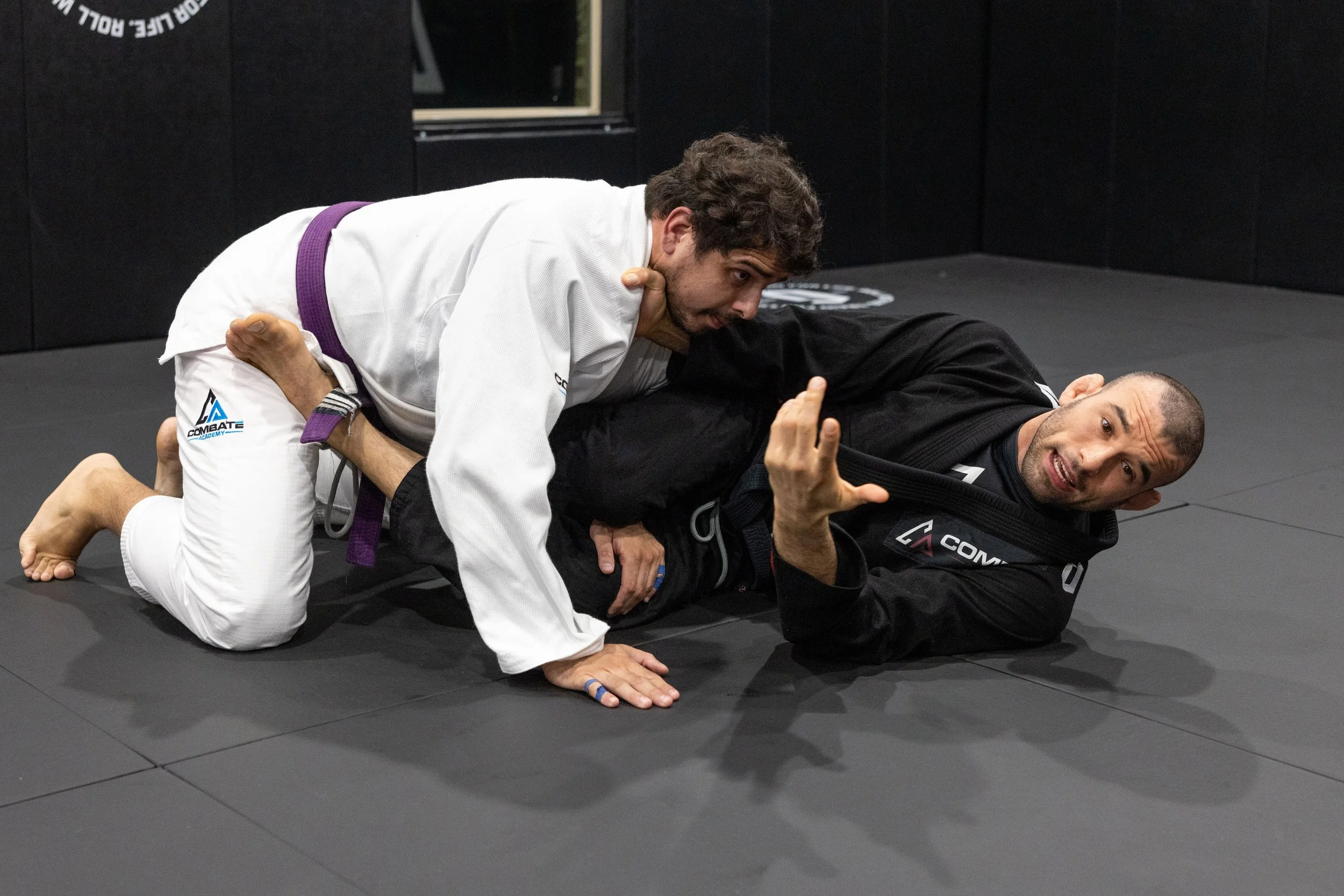Two men practicing Brazilian Jiu-Jitsu on a black mat inside a gym. One man in a white gi with a purple belt is on top, applying a submission hold on the other man in a black gi.
