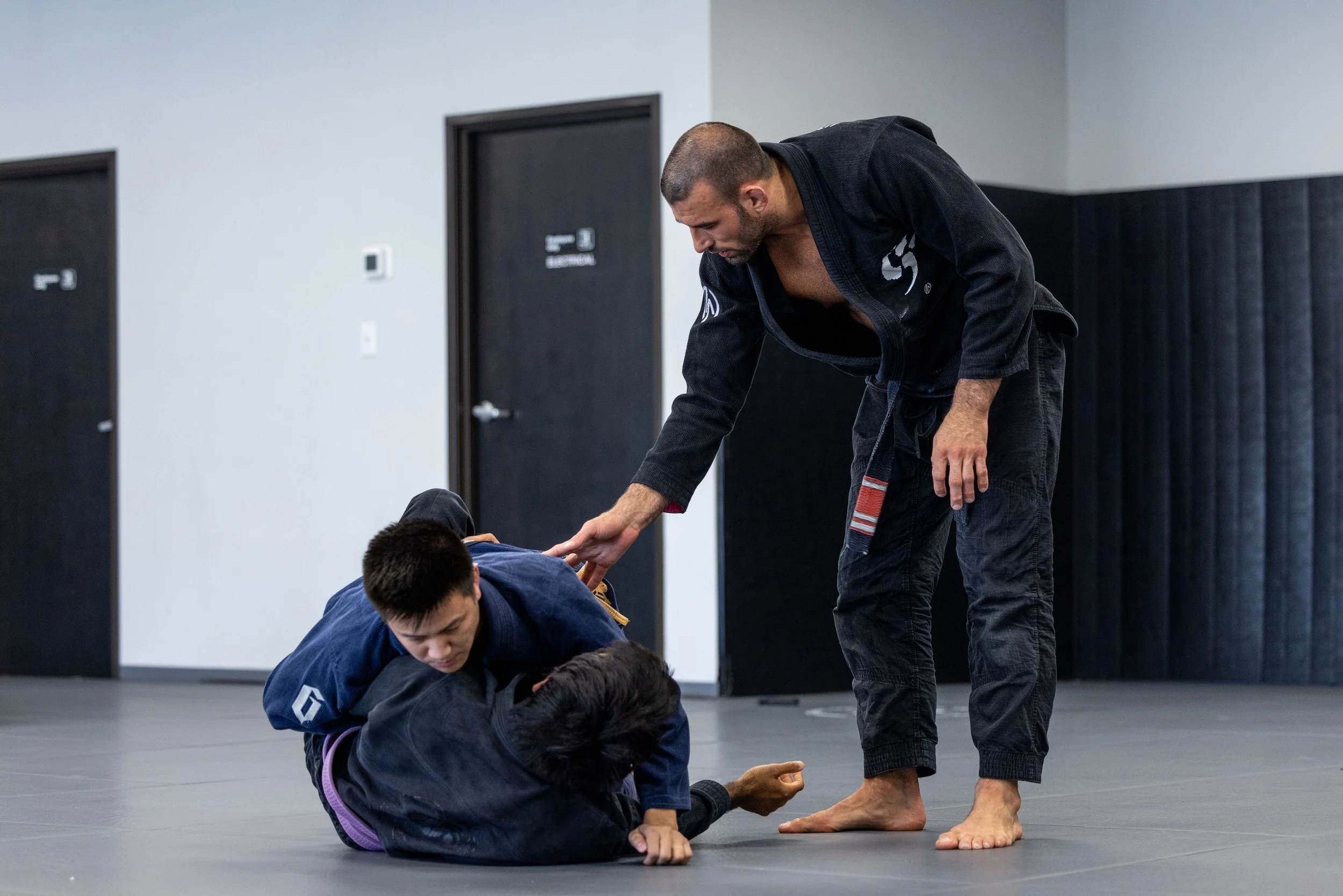 A martial arts instructor, dressed in a black gi, demonstrates a technique on two students on a gray mat in a dojo. One student, wearing a blue gi, is in a grappling position with another student, while the instructor observes closely.