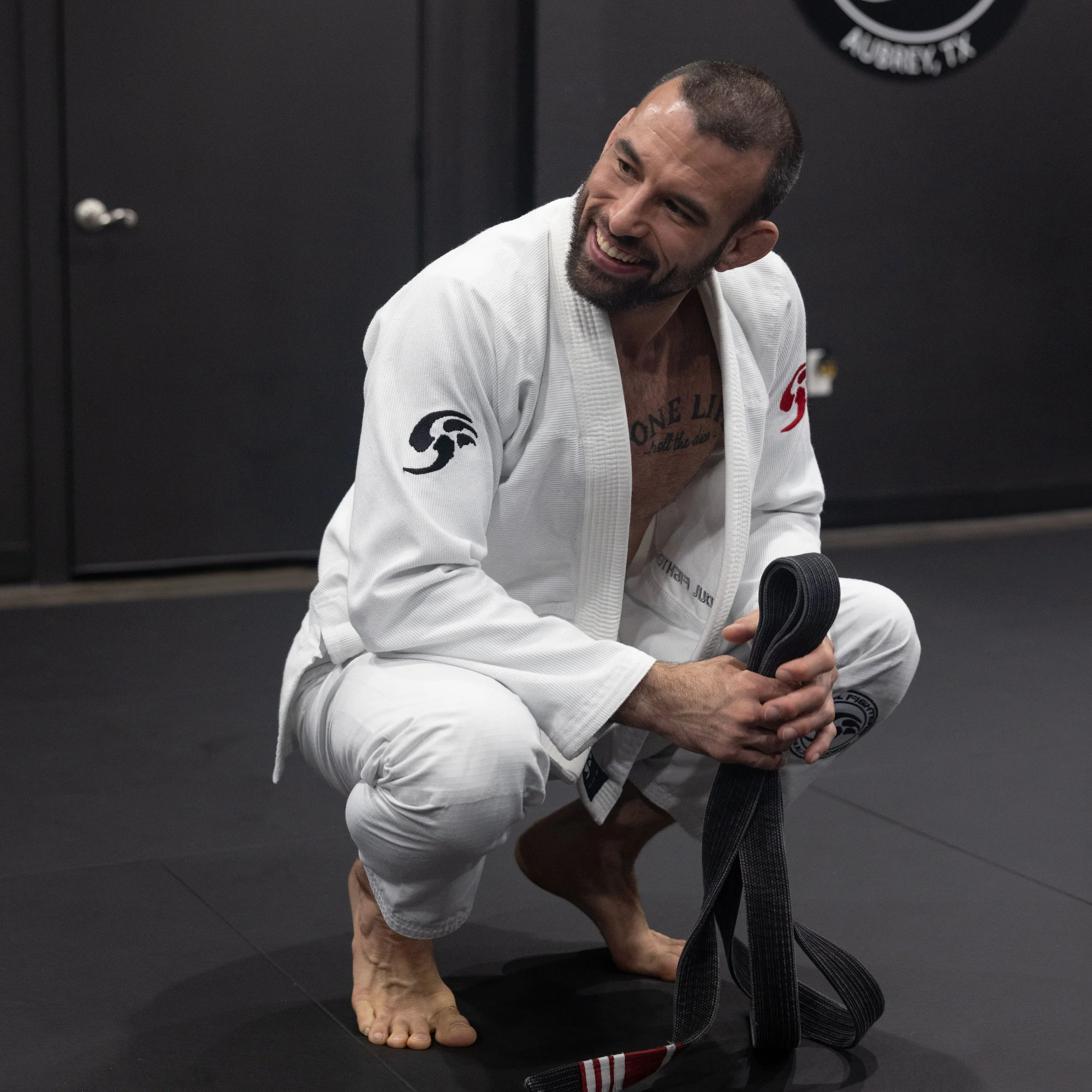 A man in a white Brazilian Jiu-Jitsu gi sits on a black mat, smiling and holding a black belt in his hand. He has short dark hair and a beard, and appears to be in a training facility.