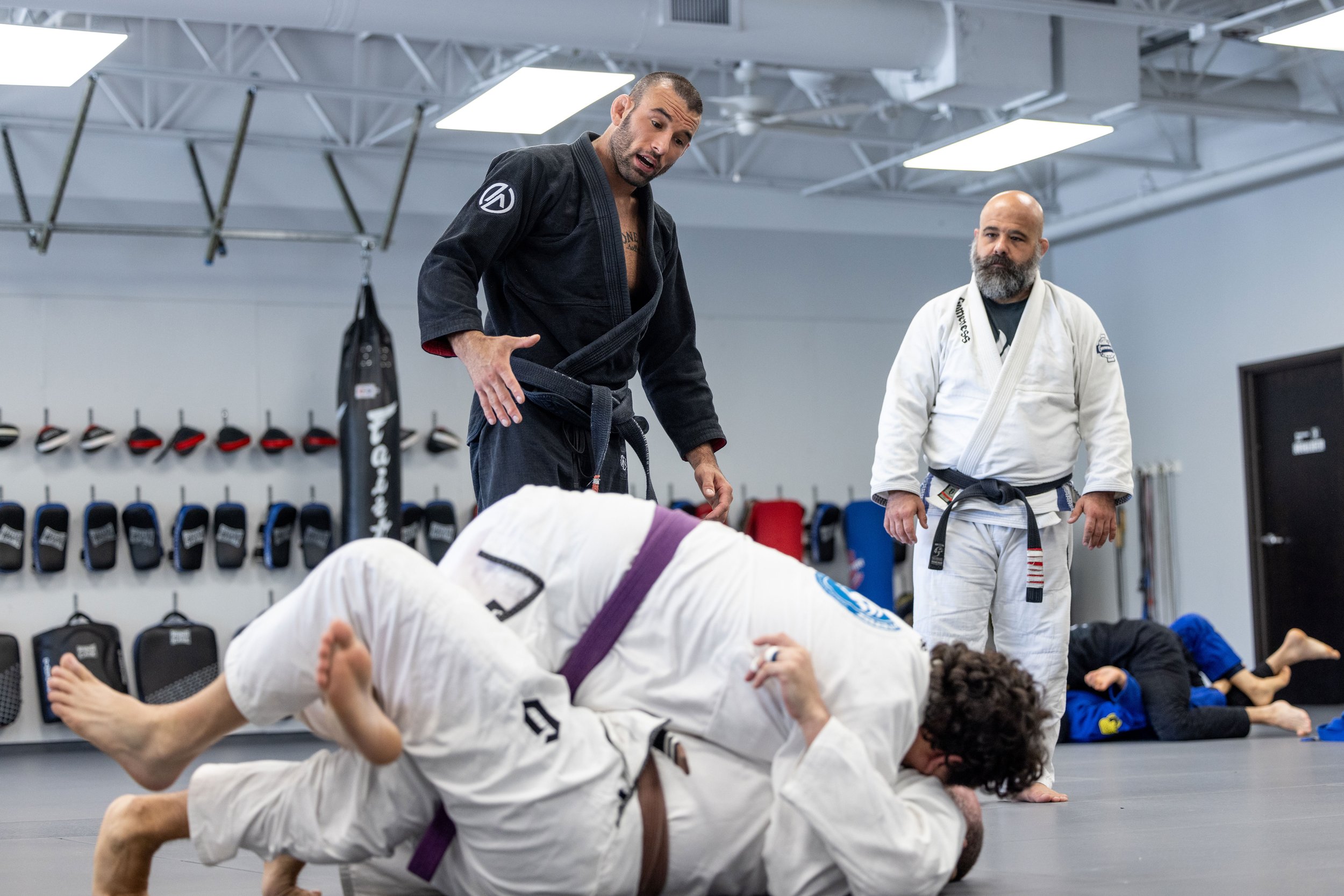 Martial arts class with students practicing Brazilian Jiu-Jitsu on mats, instructor observing, wall with hats and gloves, and students wearing gis.