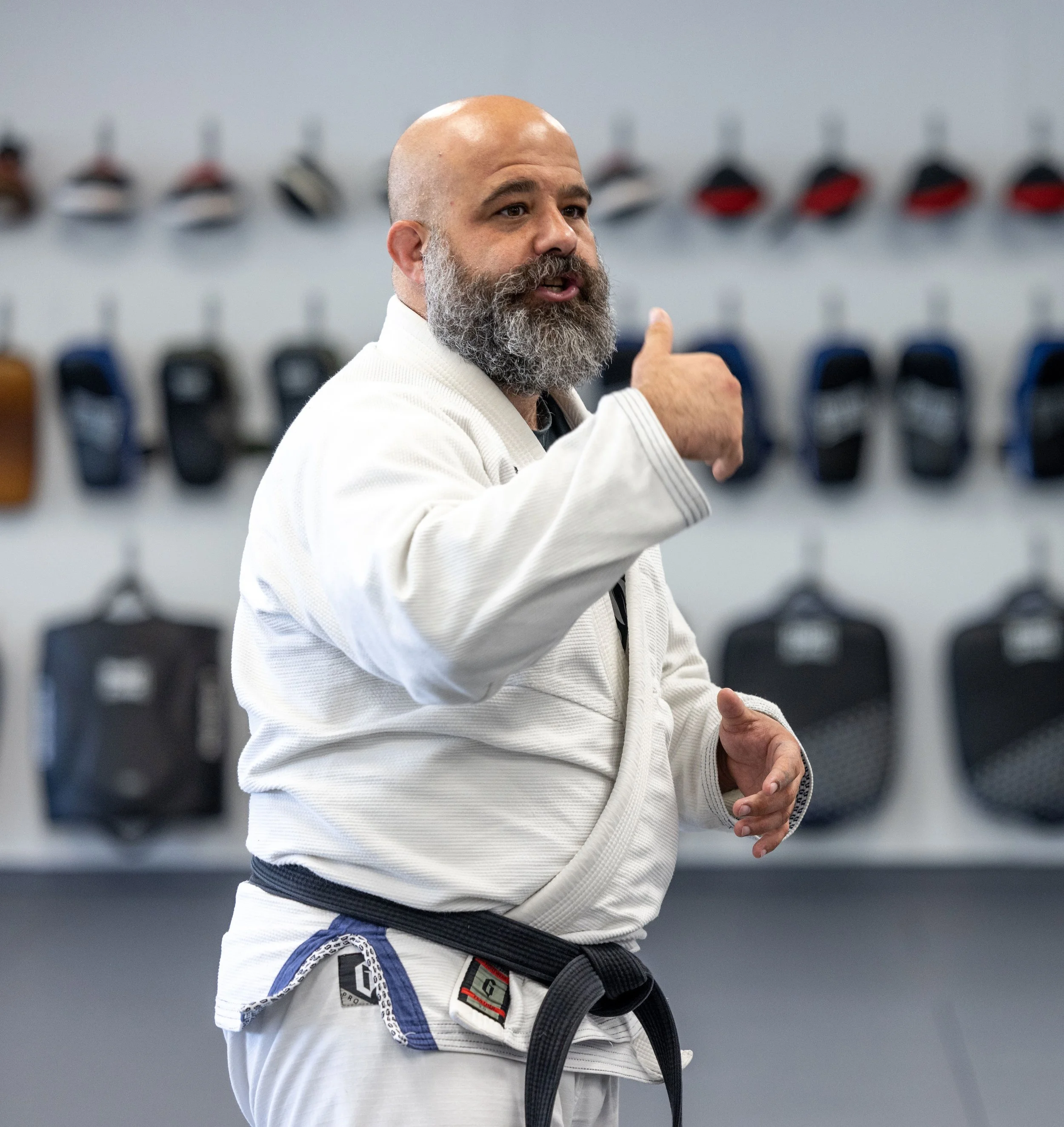 A man in a white martial arts gi with a black belt, speaking and gesturing in a martial arts training facility, with black uniforms hanging on the wall in the background.