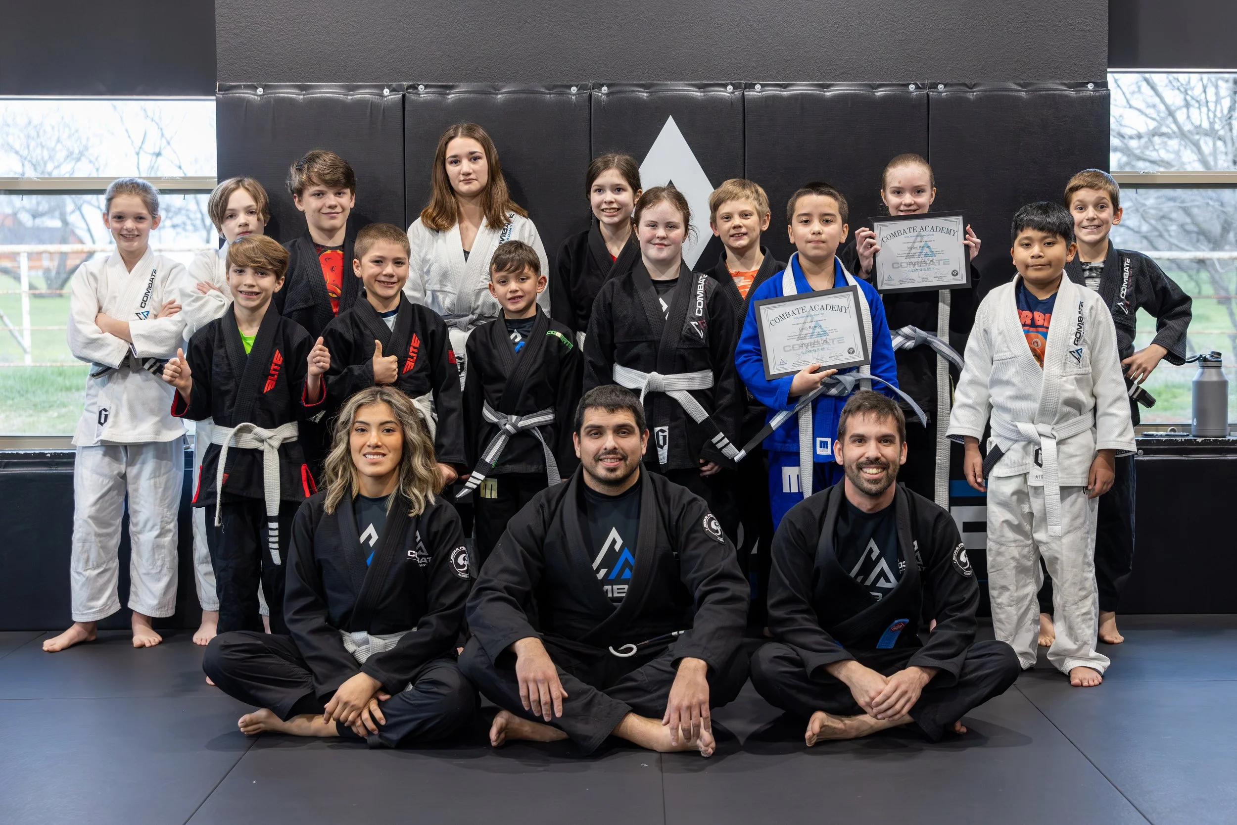 Group of children and adults in martial arts uniforms posing for a photo in a dojo, some holding certificates, with windows in the background.