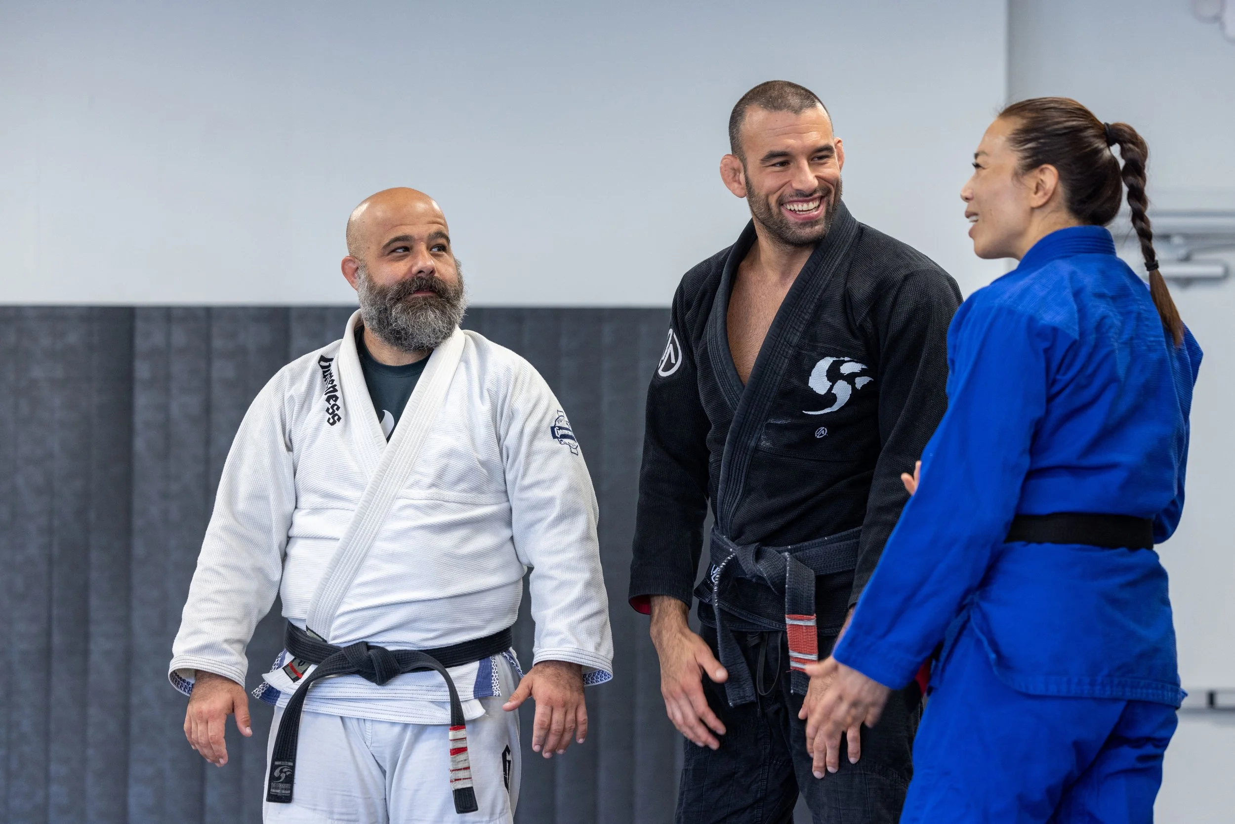 Three martial artists in gis standing in a dojo, smiling and talking.