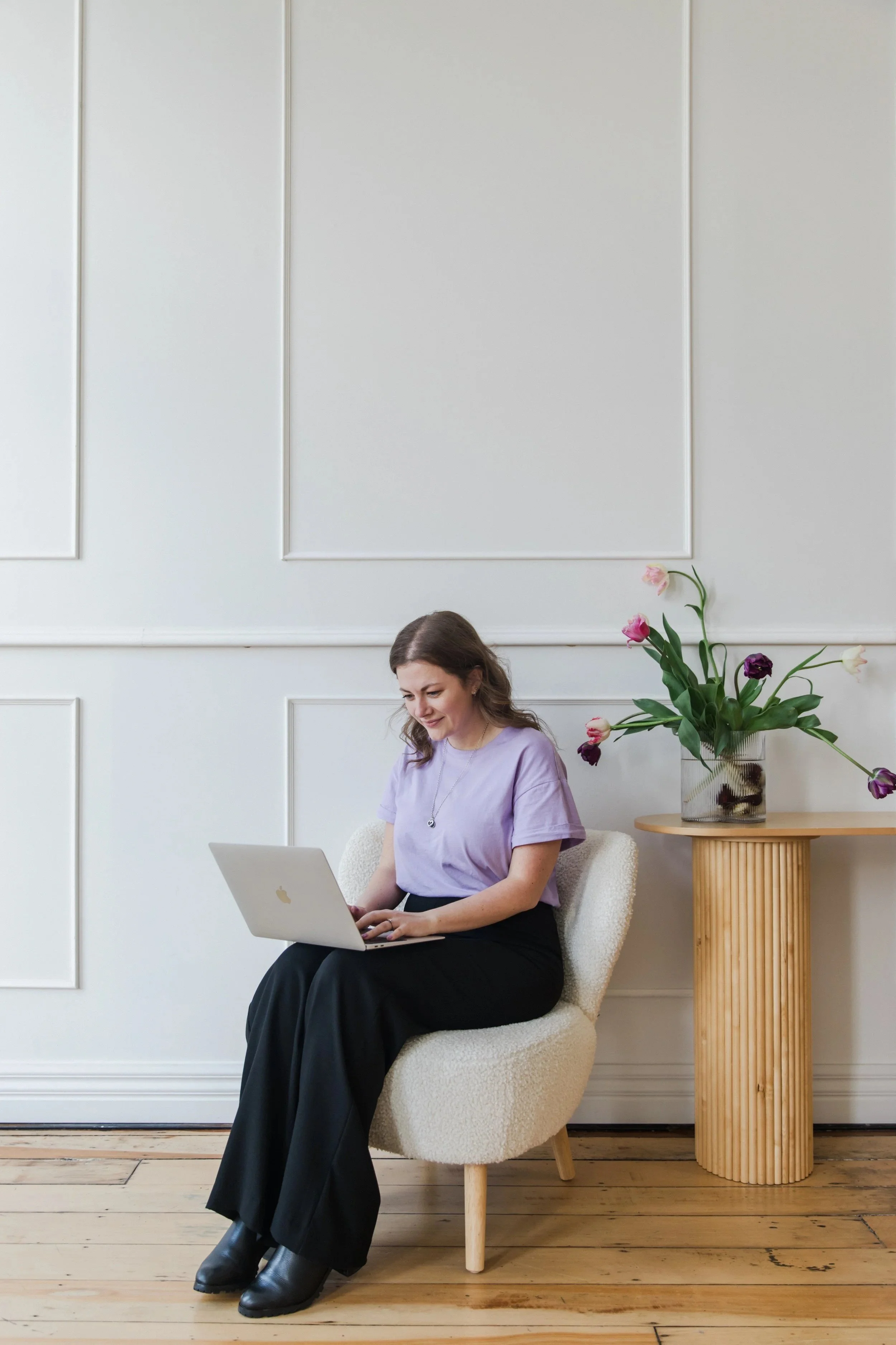 Virtual Tash - A woman sitting on a cream-colored chair, looking at her laptop with a smile, beside a wooden side table with a vase of tulips, in a white room with decorative wall panels.