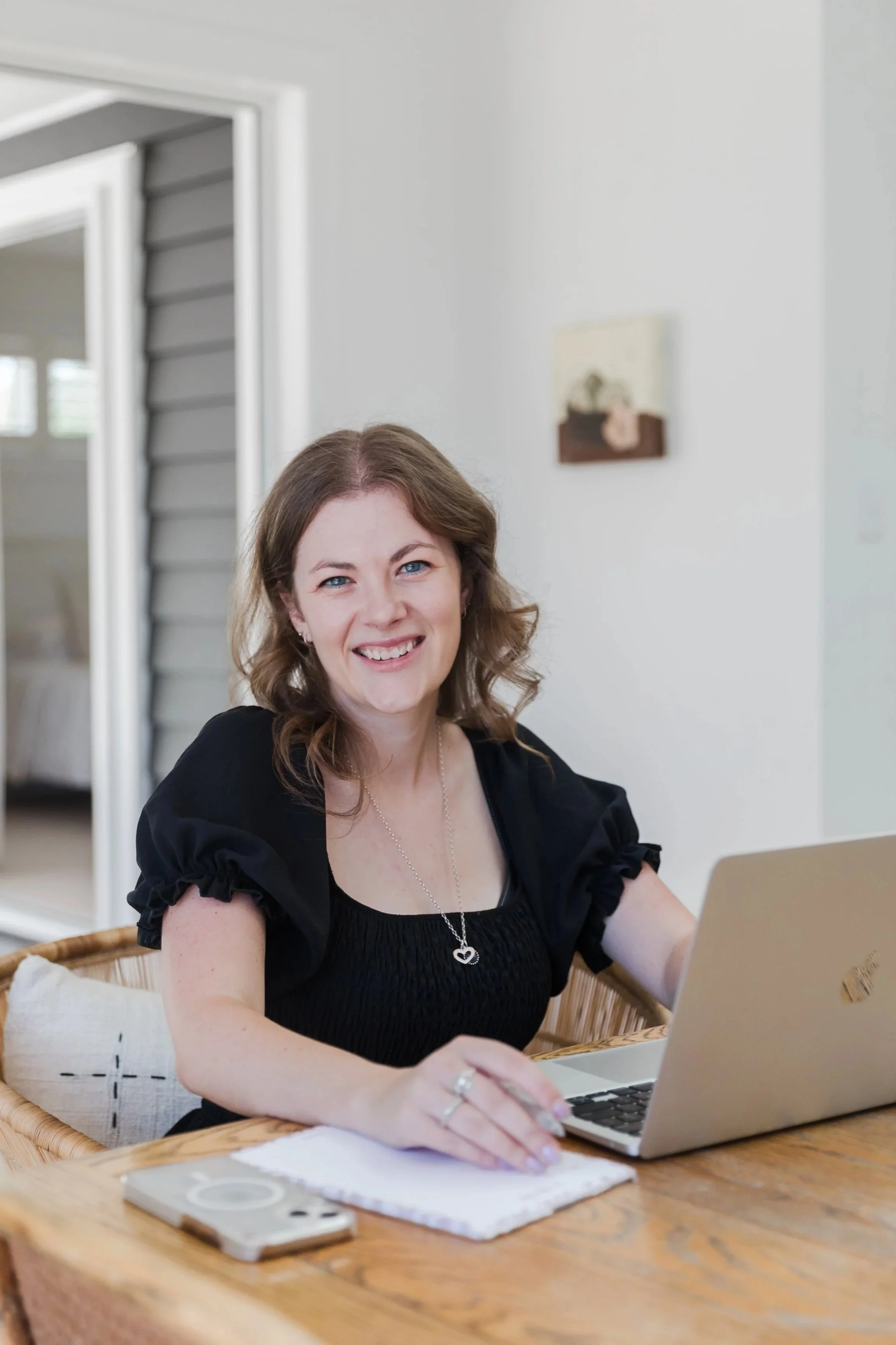 Virtual Tash - A woman with brown hair and blue eyes sitting at a wooden table, smiling, with a laptop and a notepad in front of her, in a bright room with a white wall and a painting hanging on it.