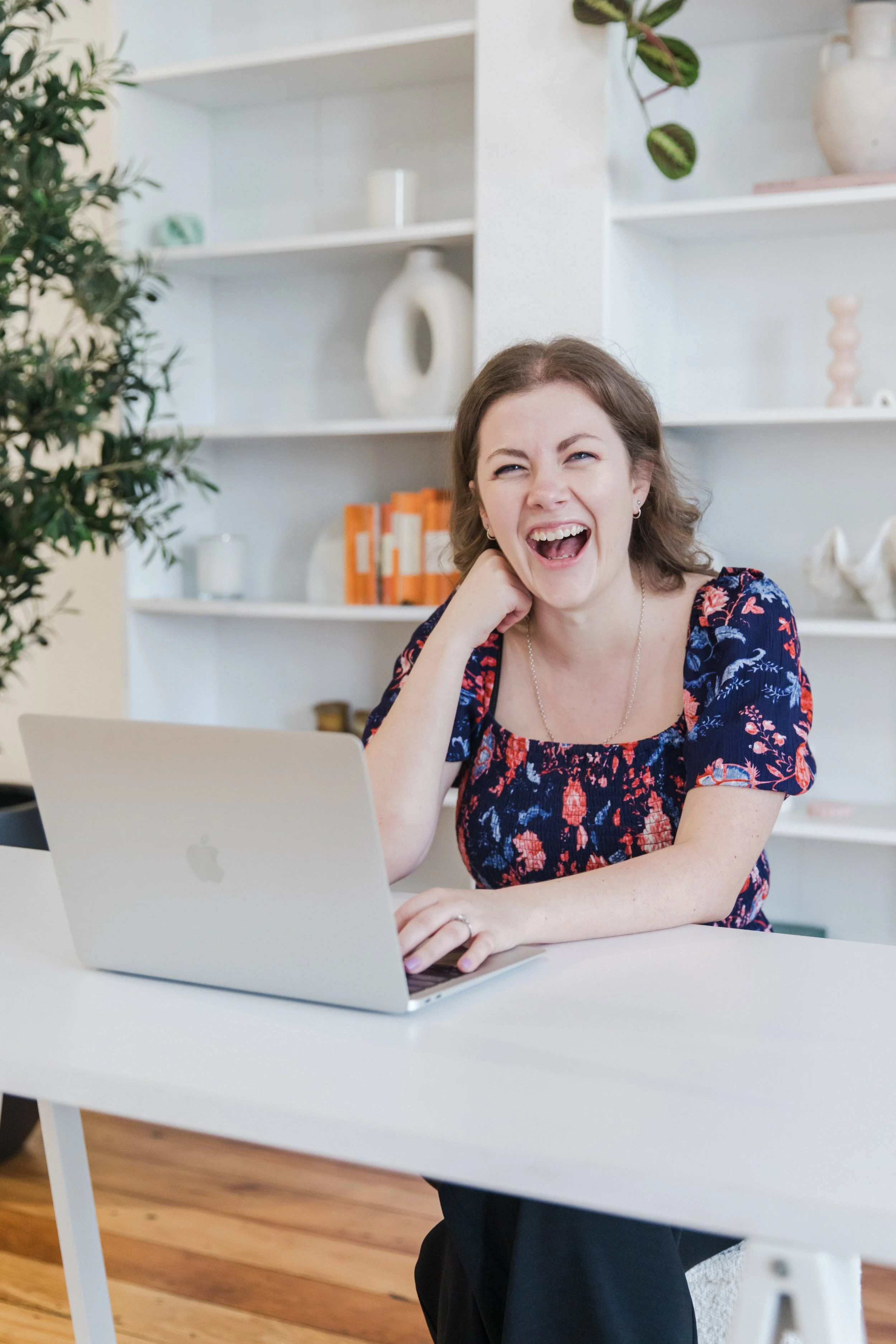Virtual Tash, Founder - sitting at a white desk with an open MacBook, smiling and laughing, in a bright room with white shelves and decorative items in the background.