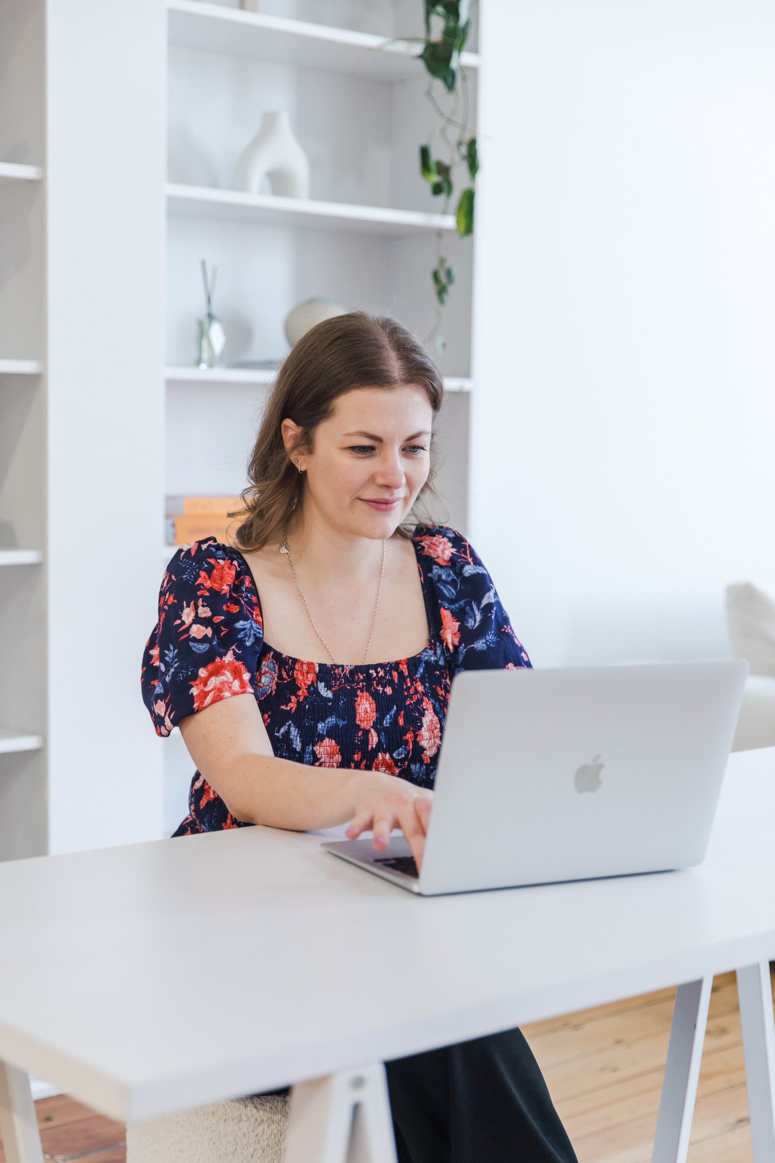 Virtual Tash, Founder - brown hair, wearing a purple floral top, sitting at a white table, working on a silver Apple MacBook Air laptop in a bright room.