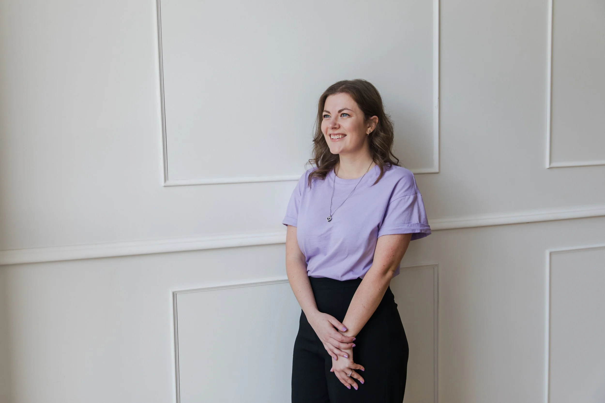 Virtual Tash, Founder, with shoulder-length brown hair wearing a lavender T-shirt and black pants, smiling and standing with hands clasped in front of her against a plain white wall.