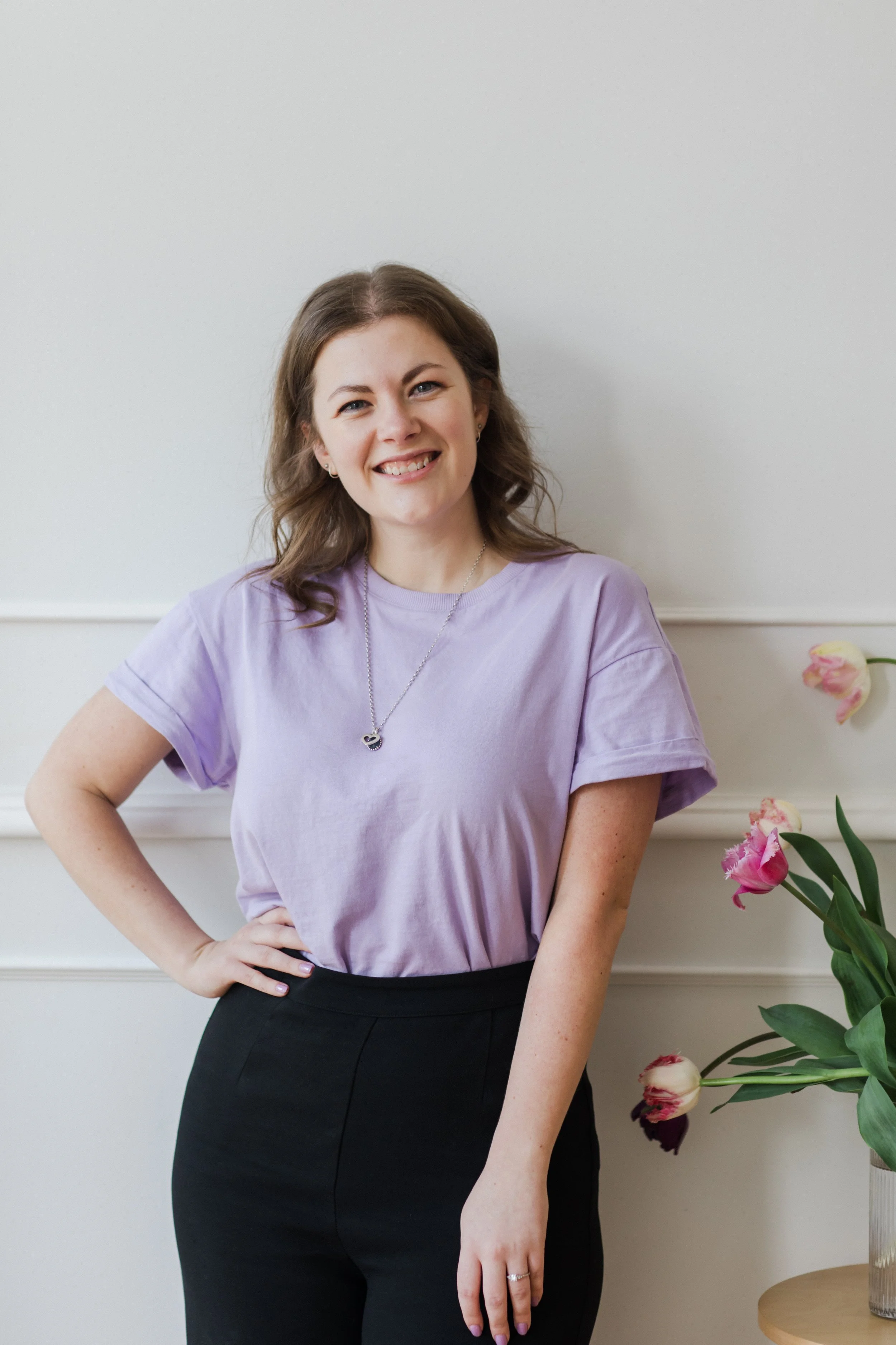 Virtual Tash, Founder - shoulder-length brown hair, wearing a lavender T-shirt, black pants, and a necklace, smiling and posing with her hand on her hip, standing next to a vase of pink and white tulips.