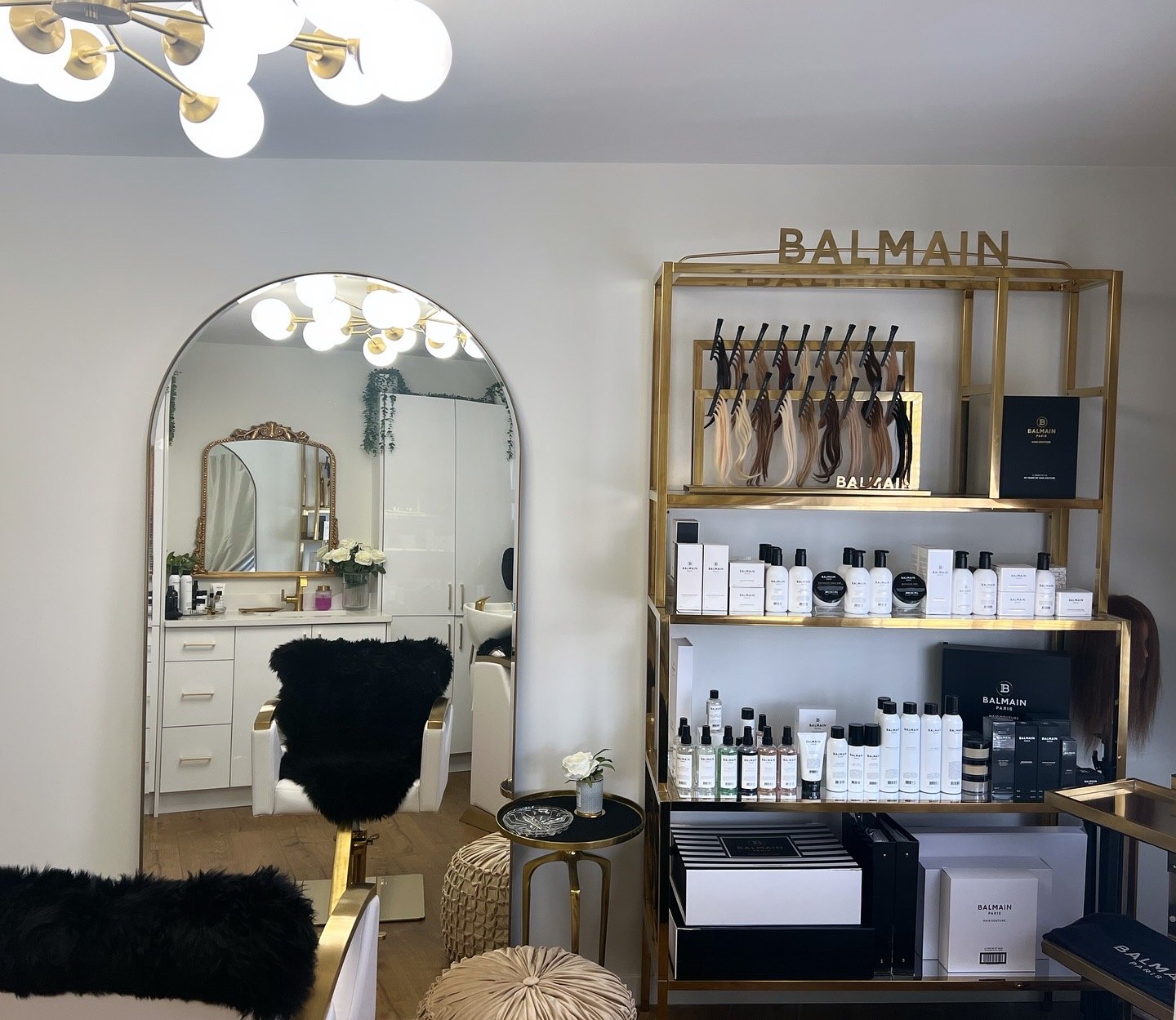 Interior of a luxury beauty or skincare boutique, featuring a large mirror with a gold frame, styling chairs with black fur covers, and a gold shelving unit displaying skincare products from Balmain, with elegant decor and lighting.