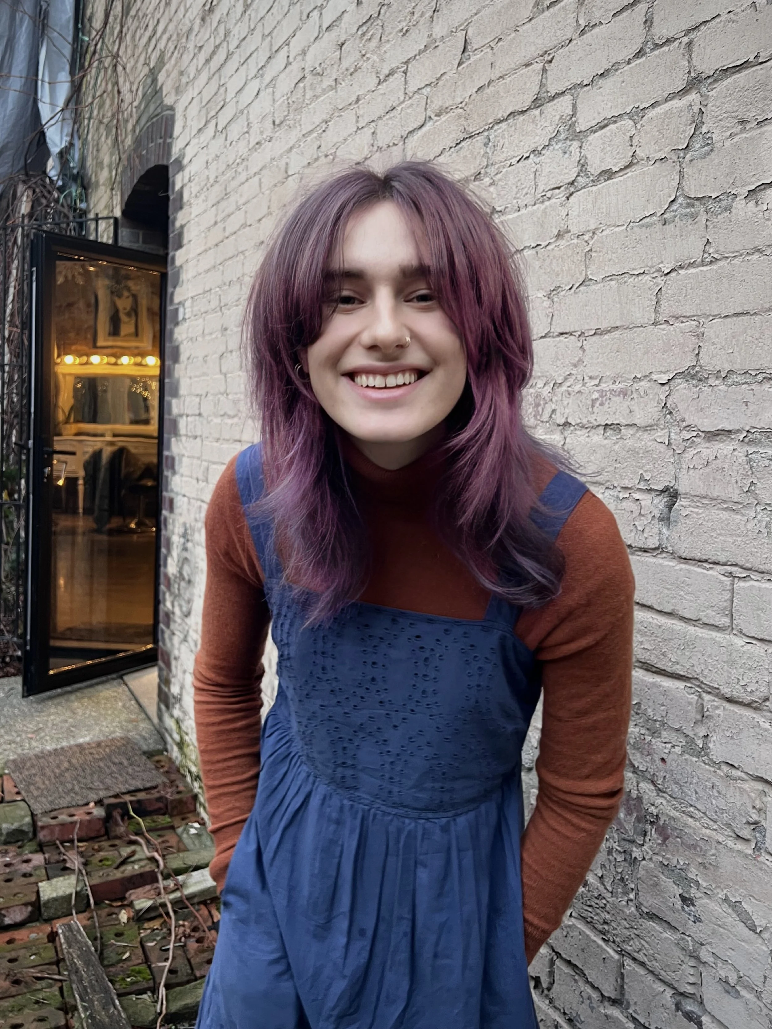Person with purple hair smiling in front of a brick wall, wearing a brown top and blue dress. Open door and decorations visible in the background.